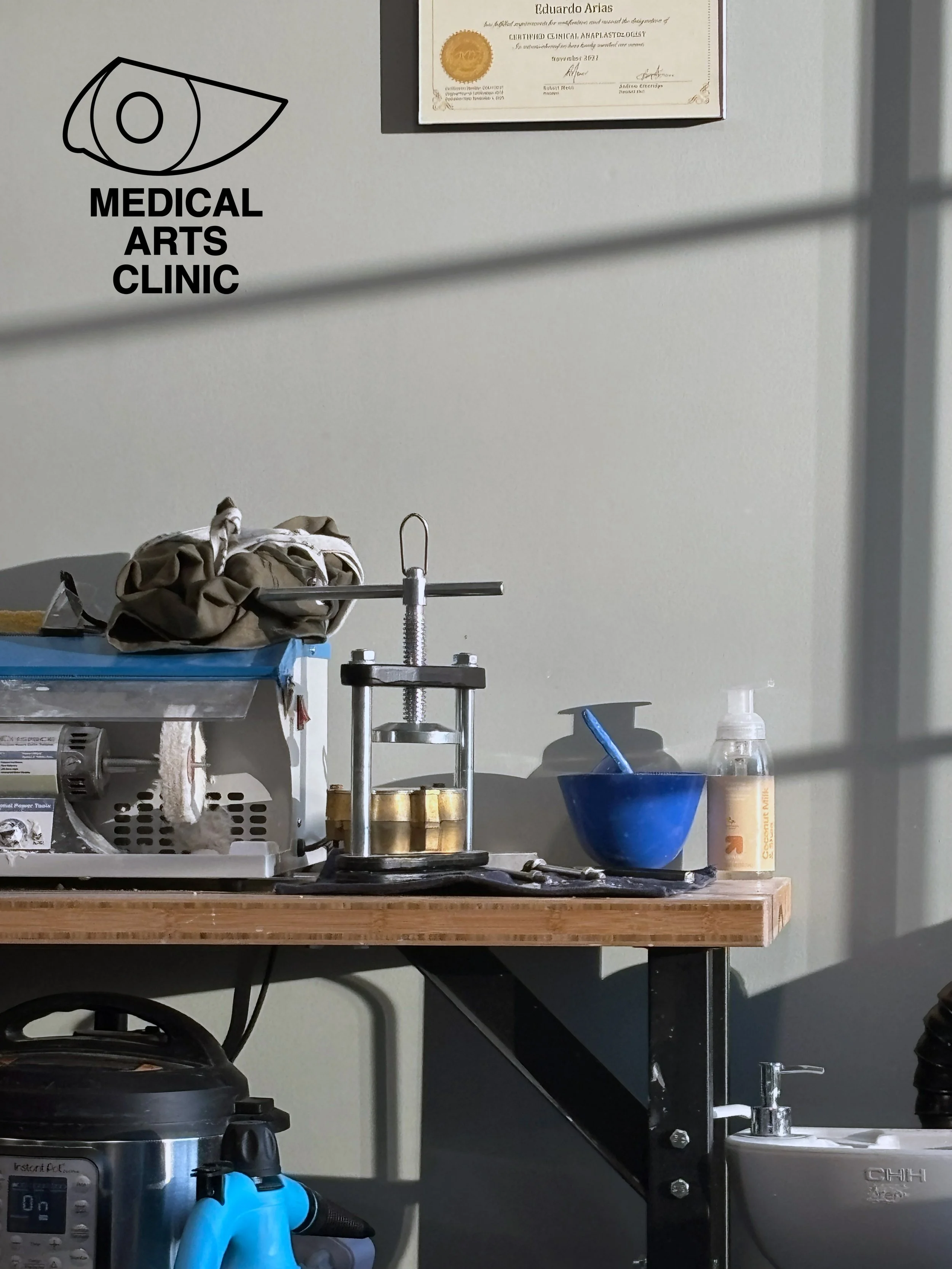 A medical workspace with a wall-mounted certificate, a logo for a medical arts clinic, and various medical and laboratory equipment on a wooden table and nearby surfaces.