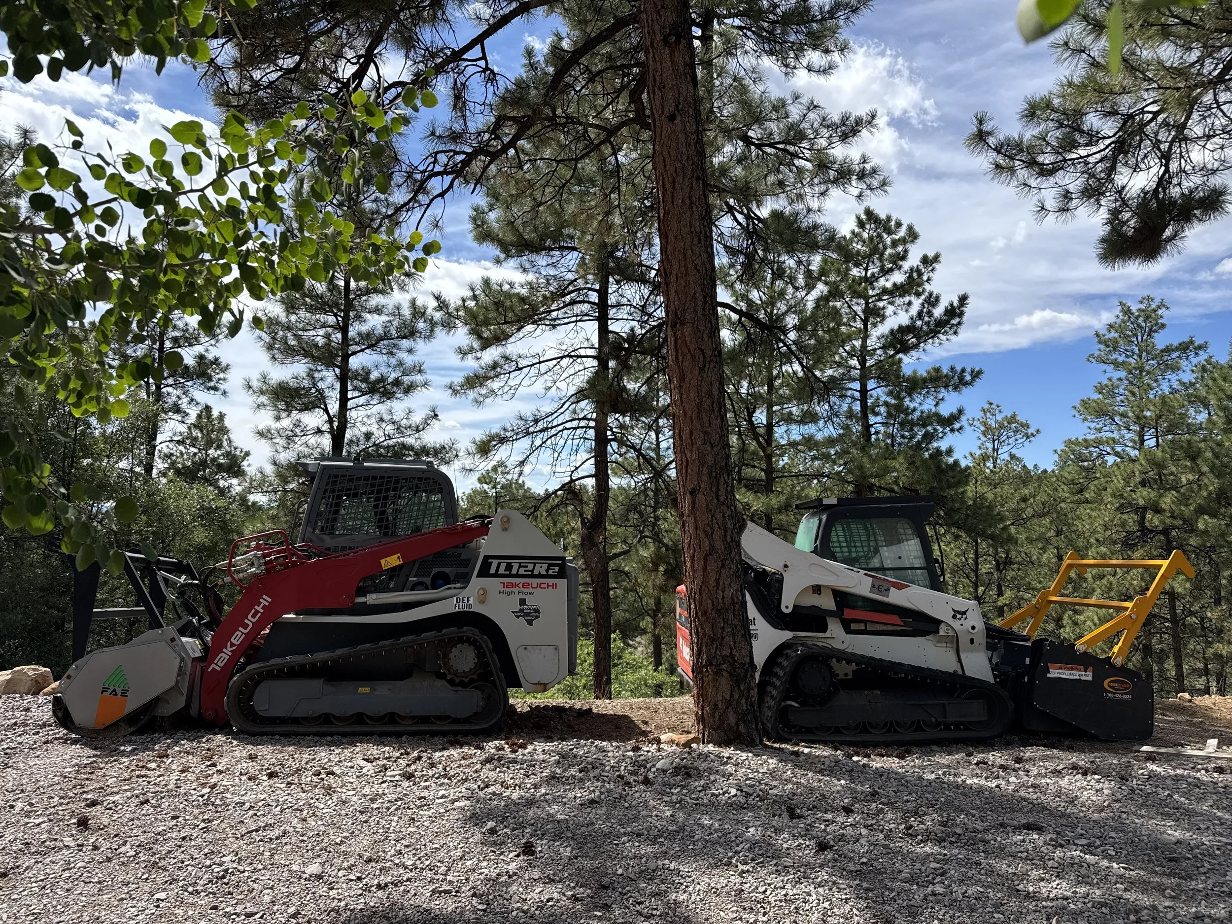Two tracked forestry mulchers parked on a dirt ground in a forested area with tall trees and a partly cloudy sky.