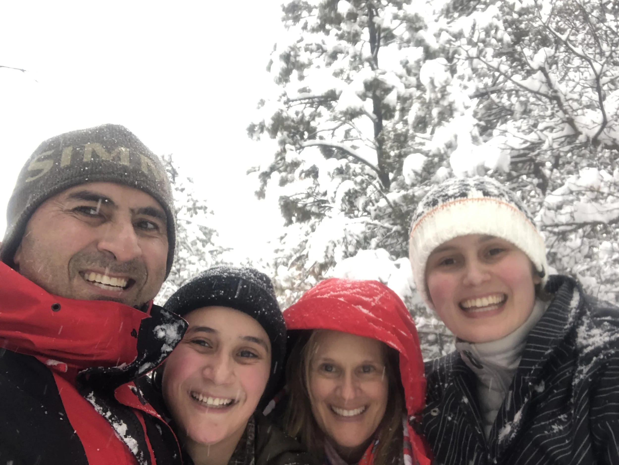 Group of smiling people taking a selfie outdoors in a snowy, wooded area, wearing winter clothing and hats.