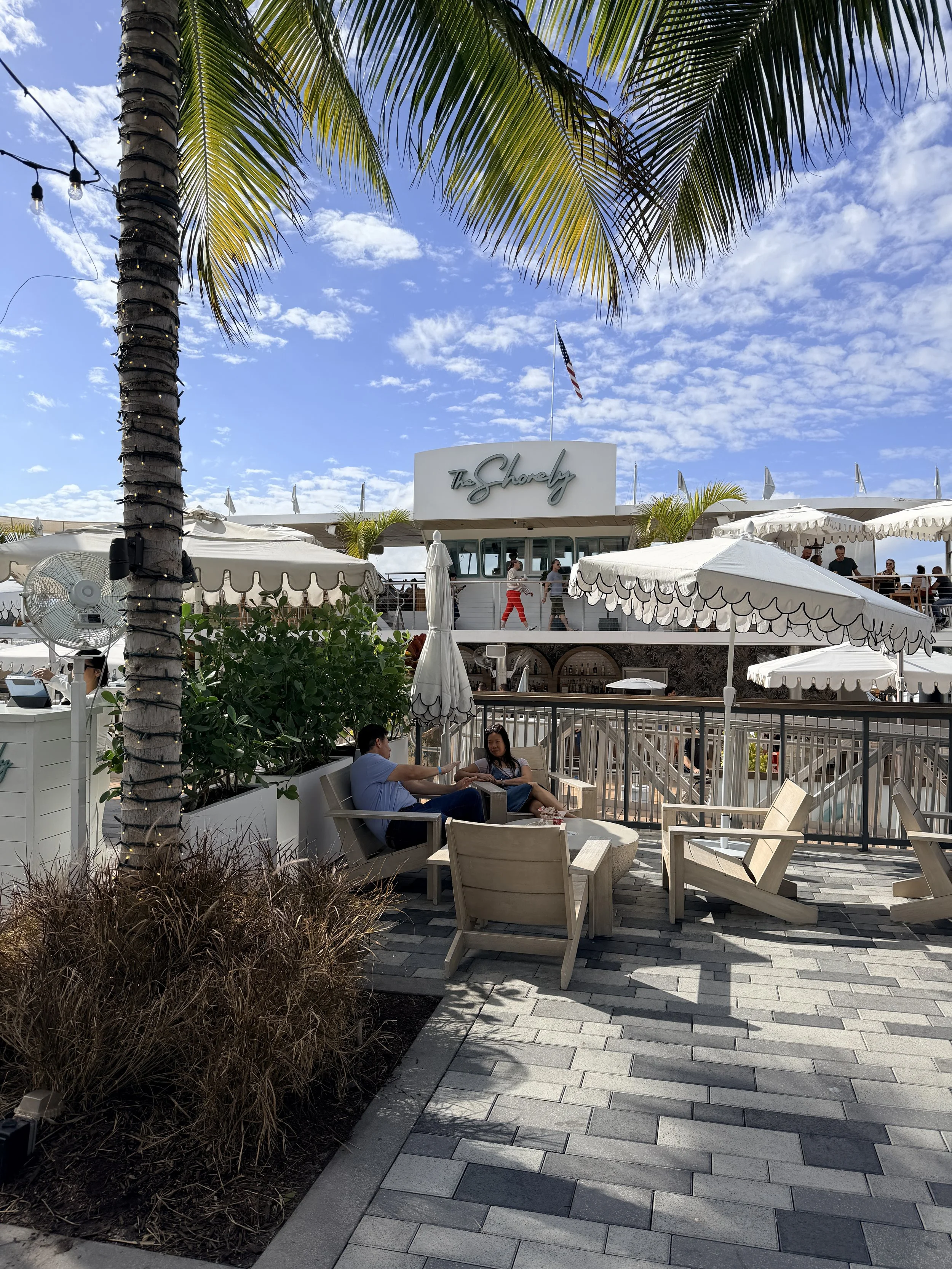 Outdoor restaurant with white umbrellas, palm trees, and a building with a sign that says "The Shorely" under a partly cloudy sky. Two people are seated on a bench in the foreground, engaging in conversation.