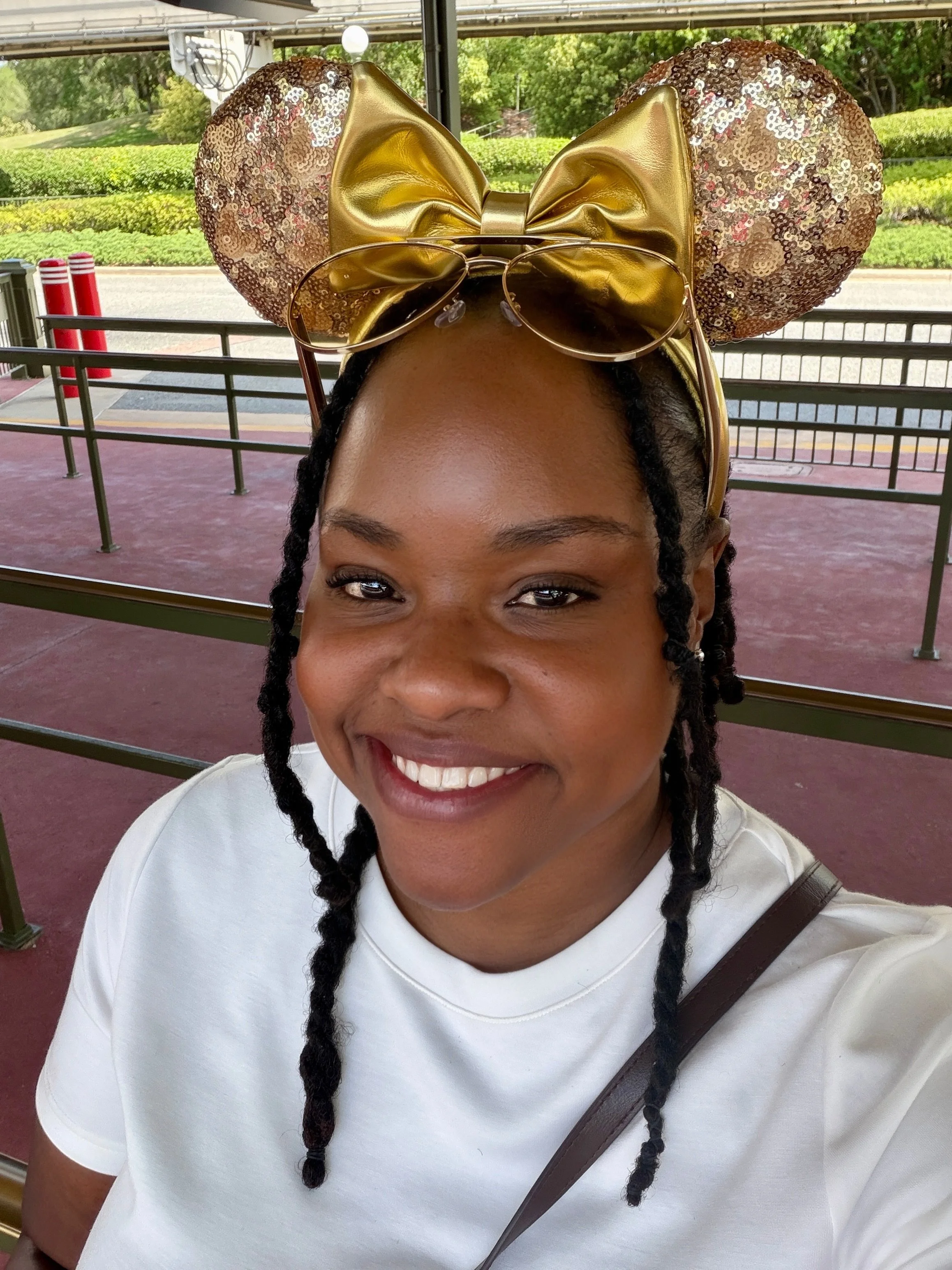 Close-up of a smiling woman with braided hair wearing a gold, sequin mouse ears headband with a large bow, sunglasses on top, and a white top, outside with green trees and a sidewalk in the background.