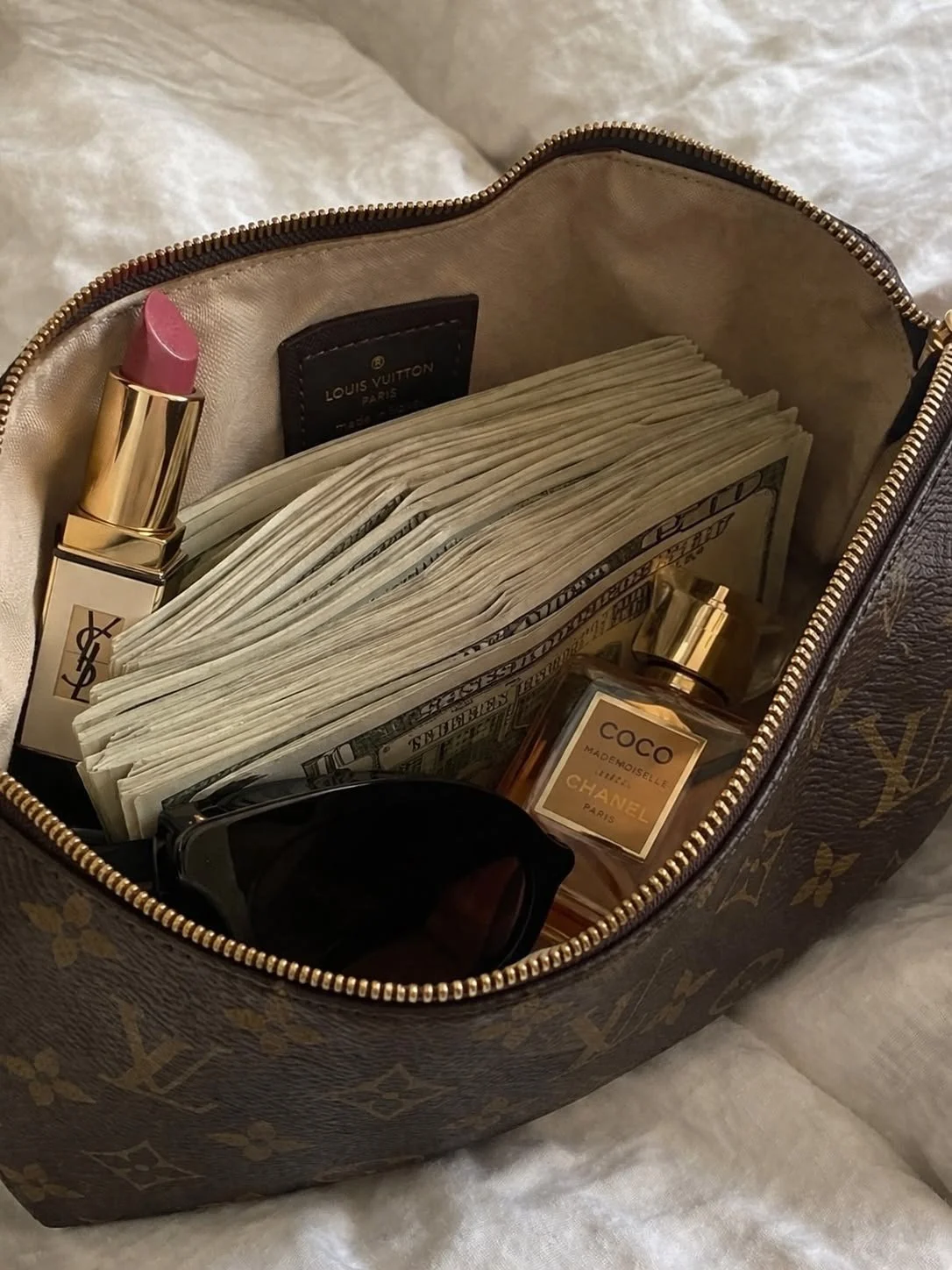 Inside a designer handbag with cash, a pink lipstick, sunglasses, a Chanel perfume, and a Louis Vuitton tag.