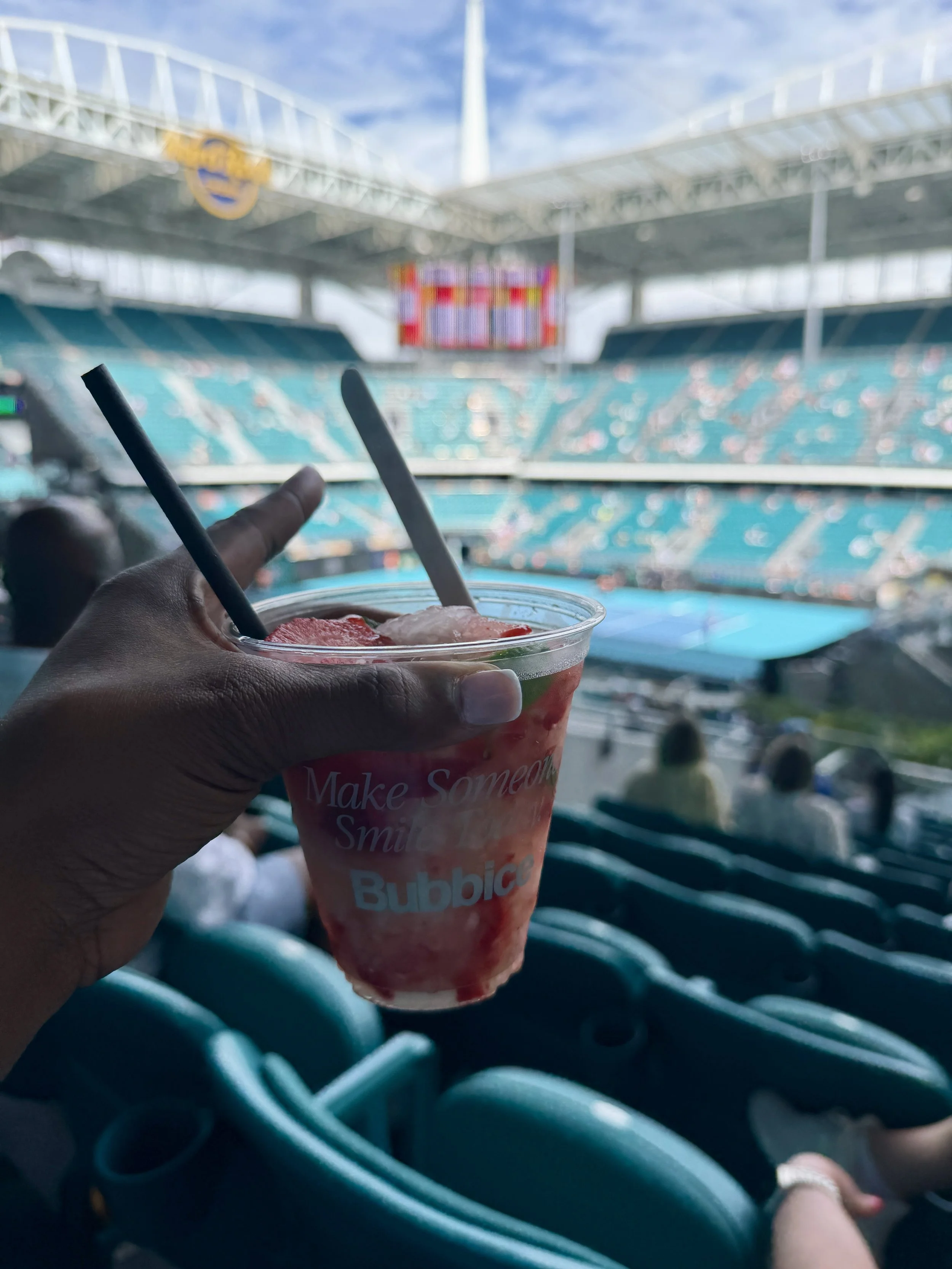 A person holding a Strawberry Lemonade bubble tea in a stadium with teal seats, a large screen, and a partly cloudy sky overhead.