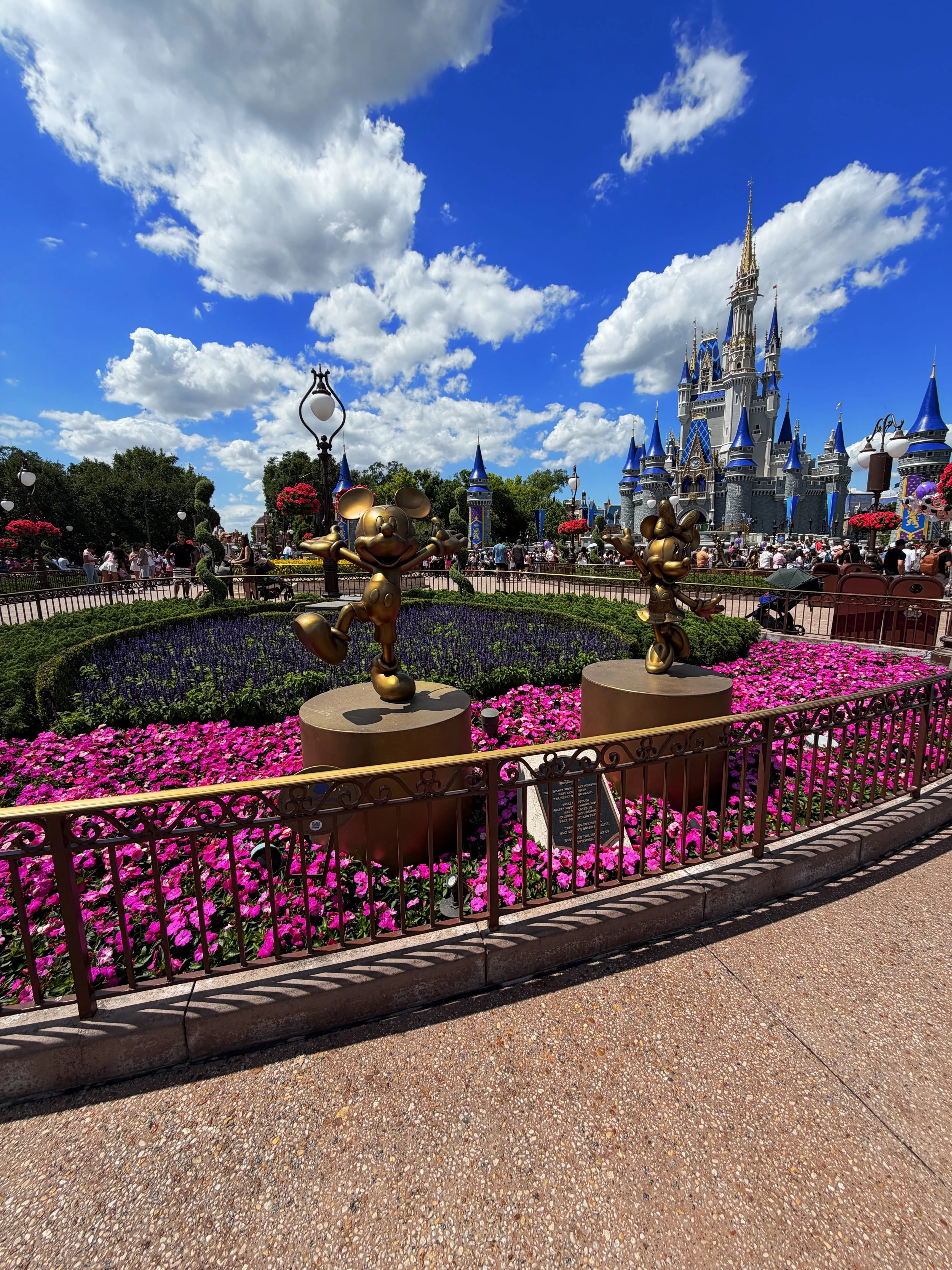 Sleeping Beauty Castle at Disneyland with Mickey Mouse statues, flower beds, and visitors walking in the park under a partly cloudy sky.