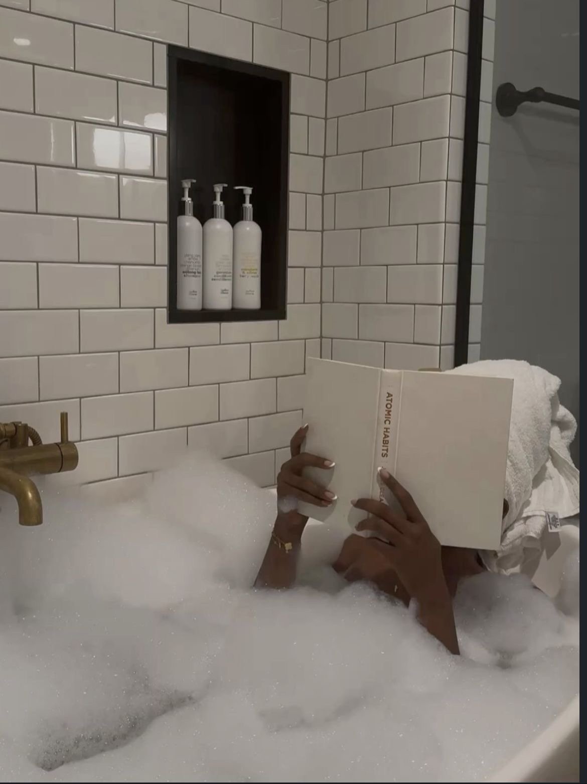 A person reading a book titled 'Atomic Habits' while taking a bubble bath, with towels in the background and bathroom fixtures visible.
