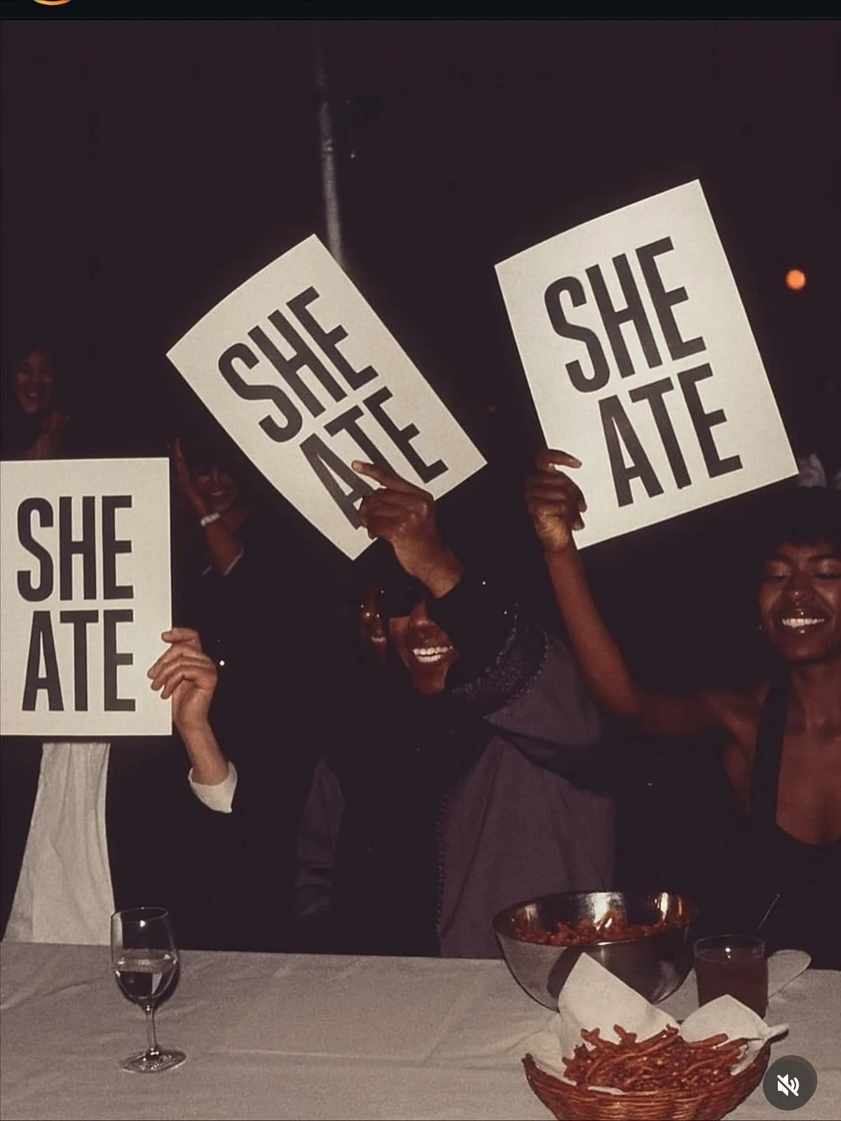 Group of six women holding signs that say 'SHE ATE' at a celebration or party, with a table in front of them containing a bowl of spaghetti and a glass of water.