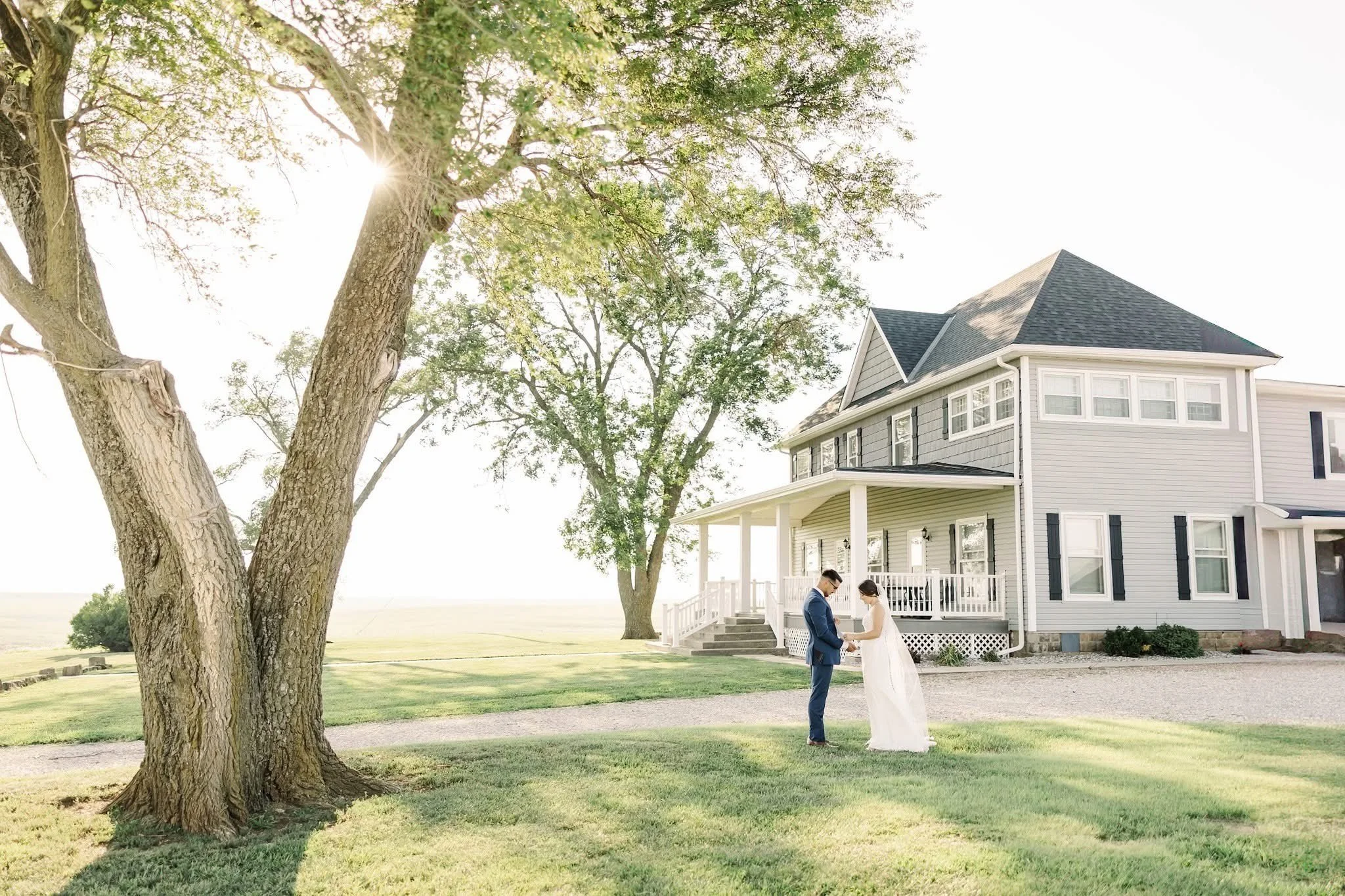 A bride and groom exchanging vows outside a large gray and white house during daytime. The scene is framed by large trees with sunlight filtering through the leaves.