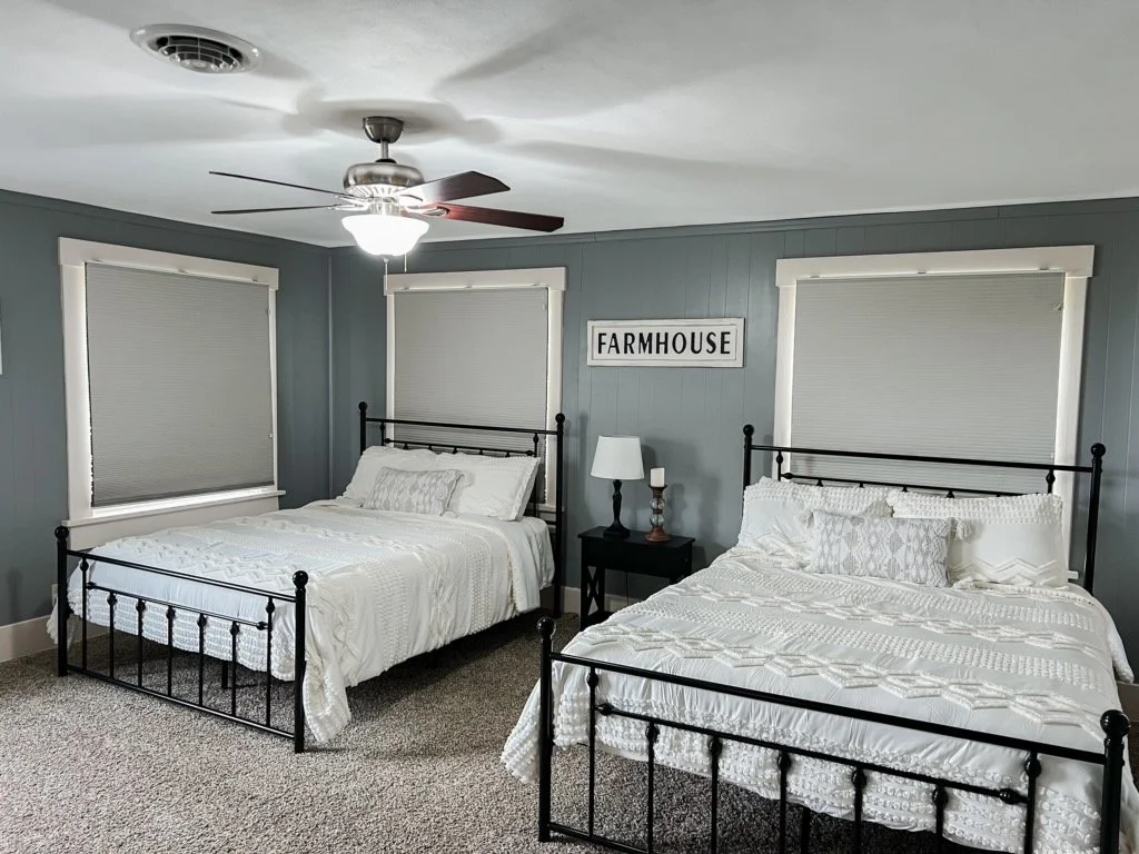 Two beds with white bedding and decorative pillows in a bedroom with gray walls, white window frames, a ceiling fan, and a sign that reads 'FARMHOUSE' on the wall.