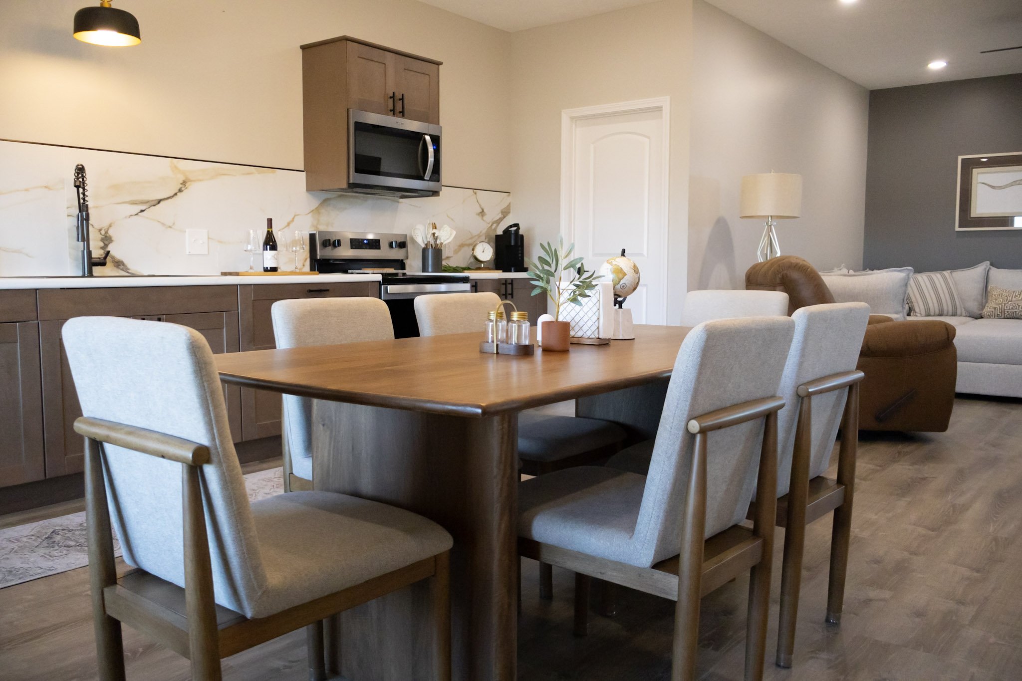 Modern kitchen and dining area with a wooden table surrounded by light upholstered chairs, a gray and white marble backsplash, stainless steel appliances, and a living space with a sofa and beige lamp.