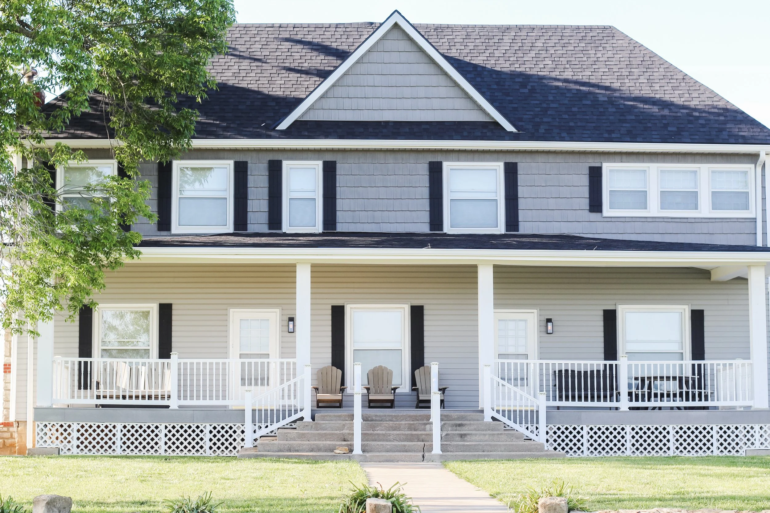 Front view of a house with a gray and white exterior, multiple windows, a porch with chairs, and a tree partially covering the house on the left side.