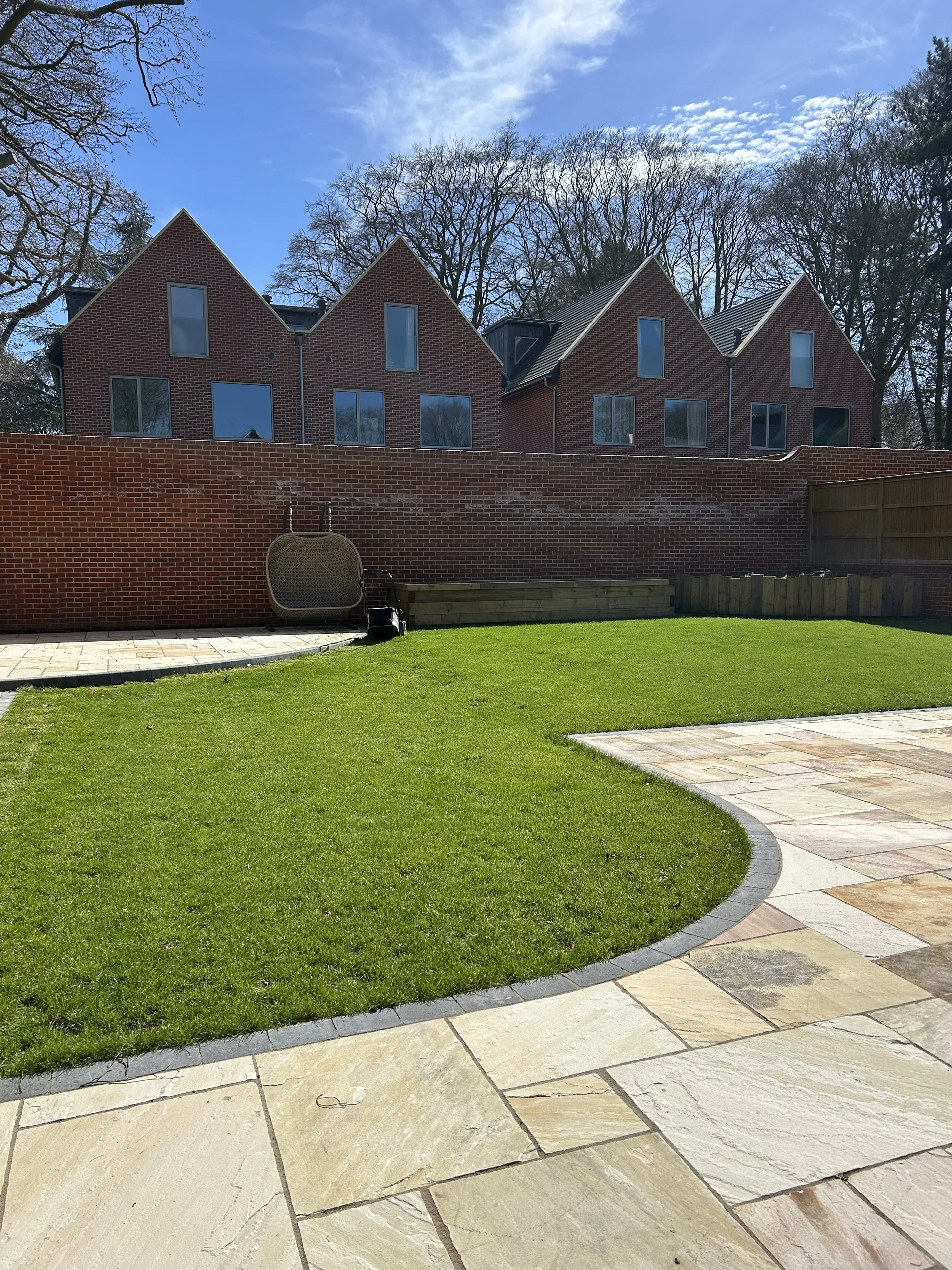 A backyard with a brick house in the distance, a green lawn, and a paved stone pathway leading up to the house. There is a wooden fence on the right side of the image and a patio area with a large outdoor chair in the foreground. Trees with no leaves are visible behind the house, with a partly cloudy sky overhead.