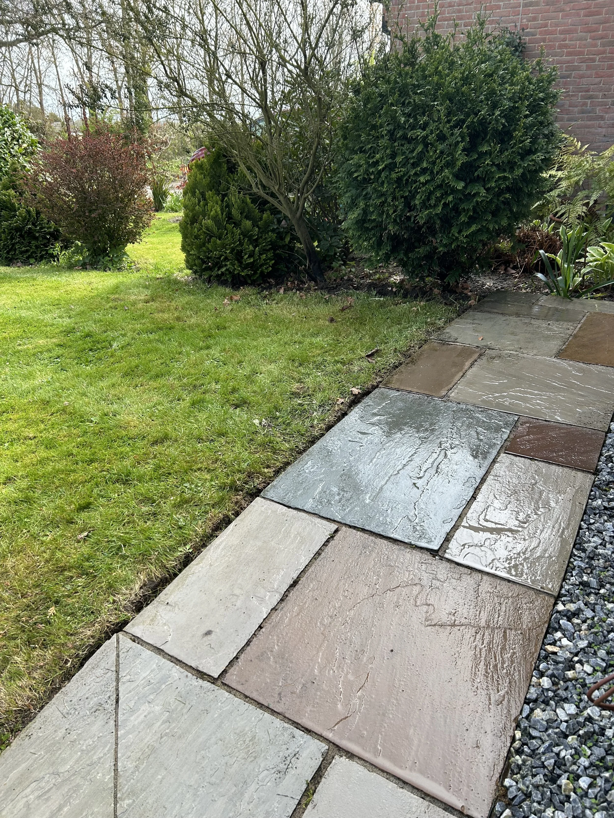 A newly cleaned and wet stone walkway with different colored concrete and stone tiles, bordered by a grass lawn and small gravel on the right side, with some green shrubs and a brick wall in the background.