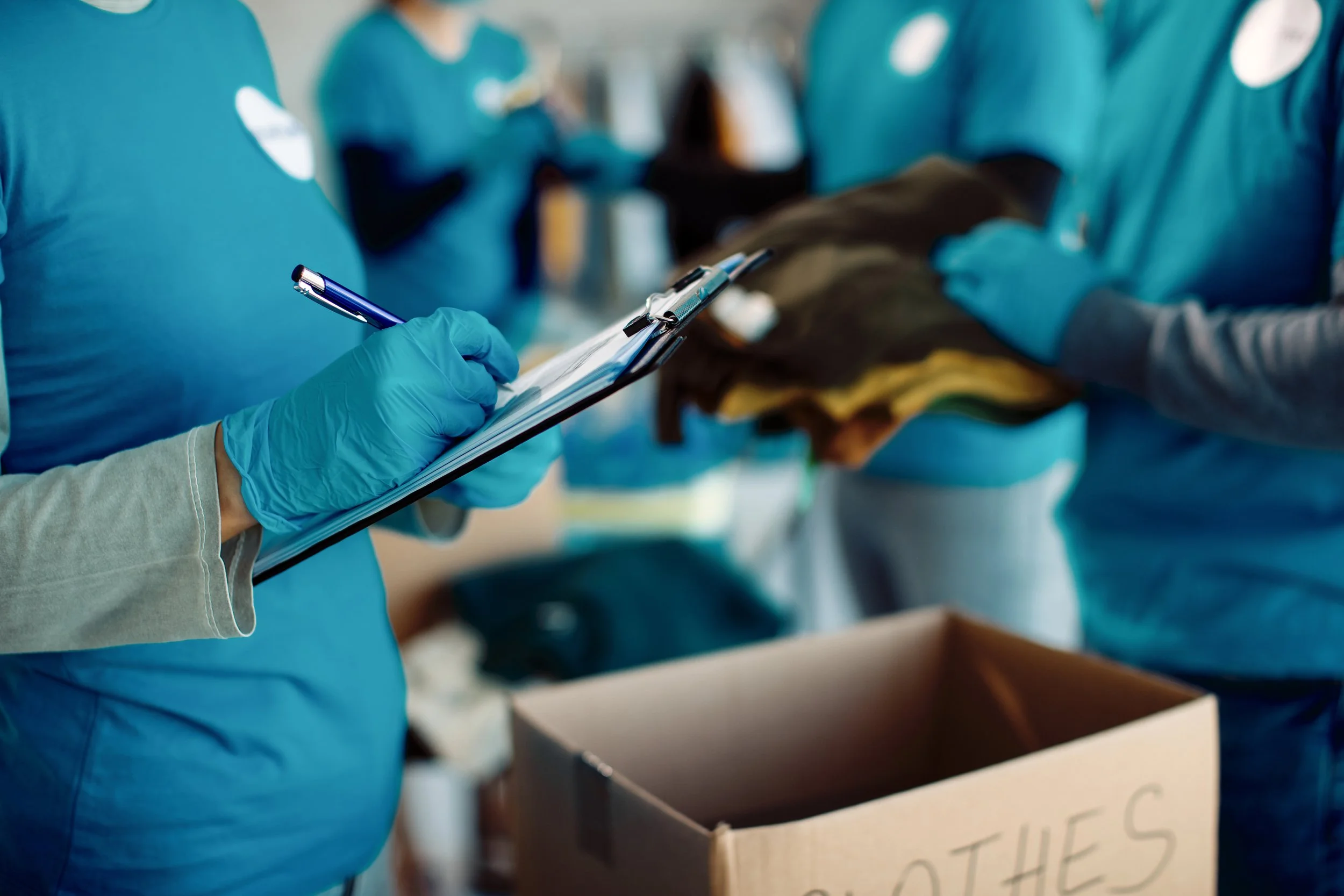 Group of healthcare workers in blue scrubs and gloves, holding clipboards, in a healthcare setting. One person is writing on a clipboard, while others are in the background.