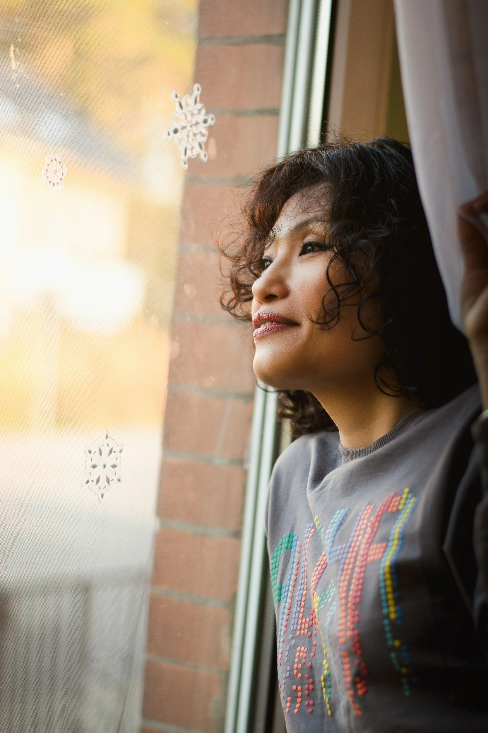 A woman with curly dark hair and a gray shirt is looking out a window with snowflake-shaped decorations on the glass.