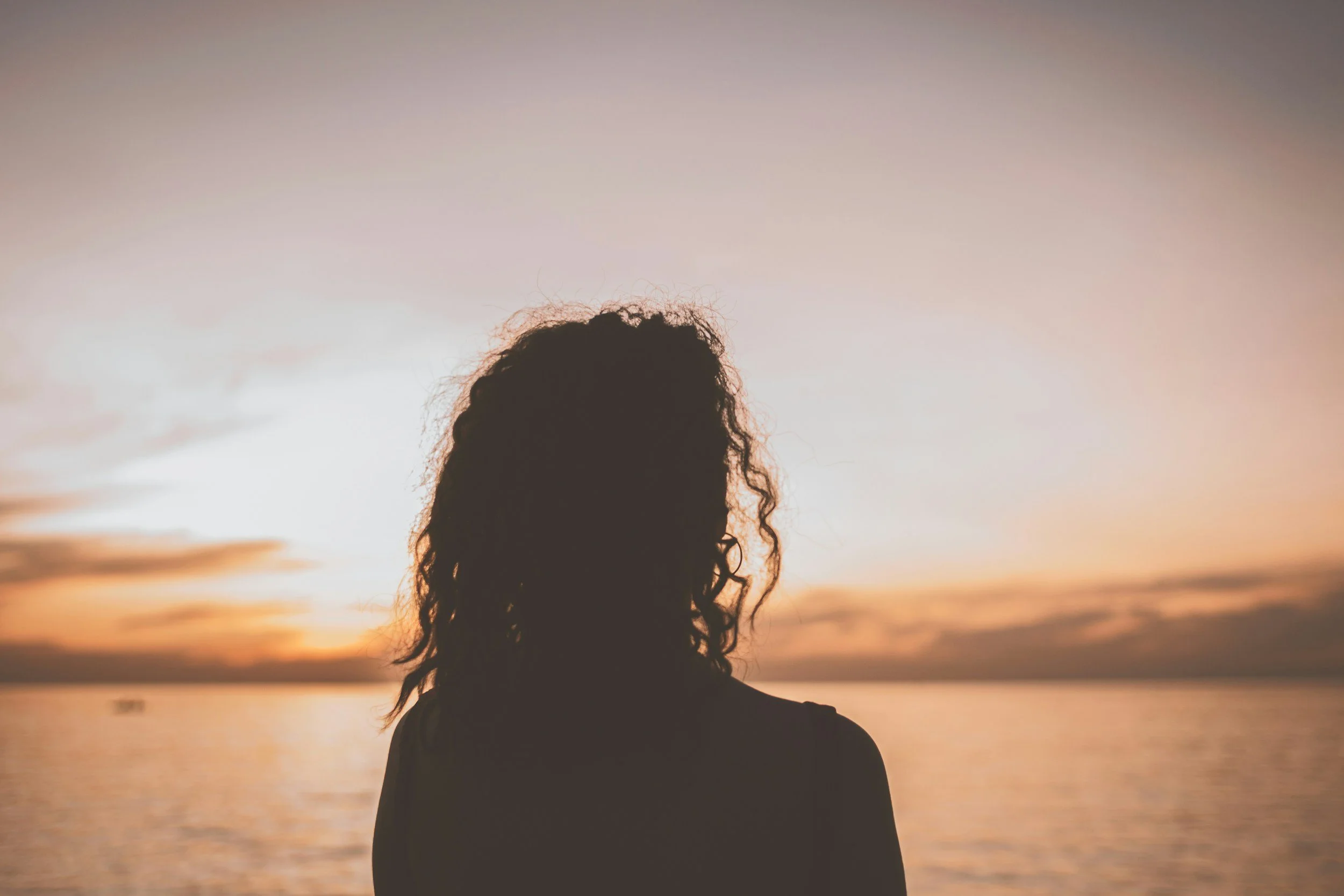 Silhouette of a woman with curly hair, standing near water during sunset or sunrise.