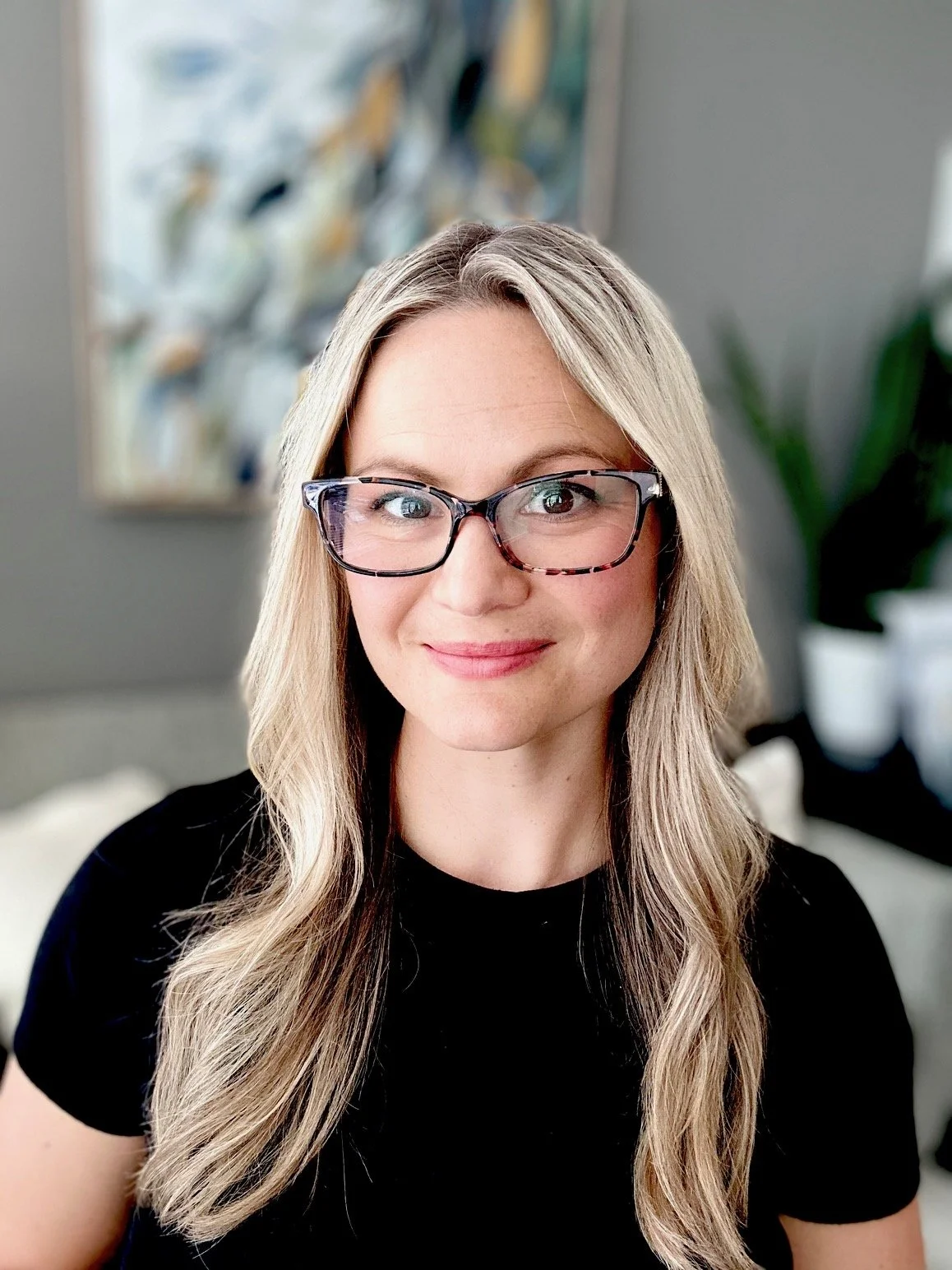 A woman with long blonde hair, wearing glasses and a black shirt, smiling indoors with a blurred artwork and plant in the background.