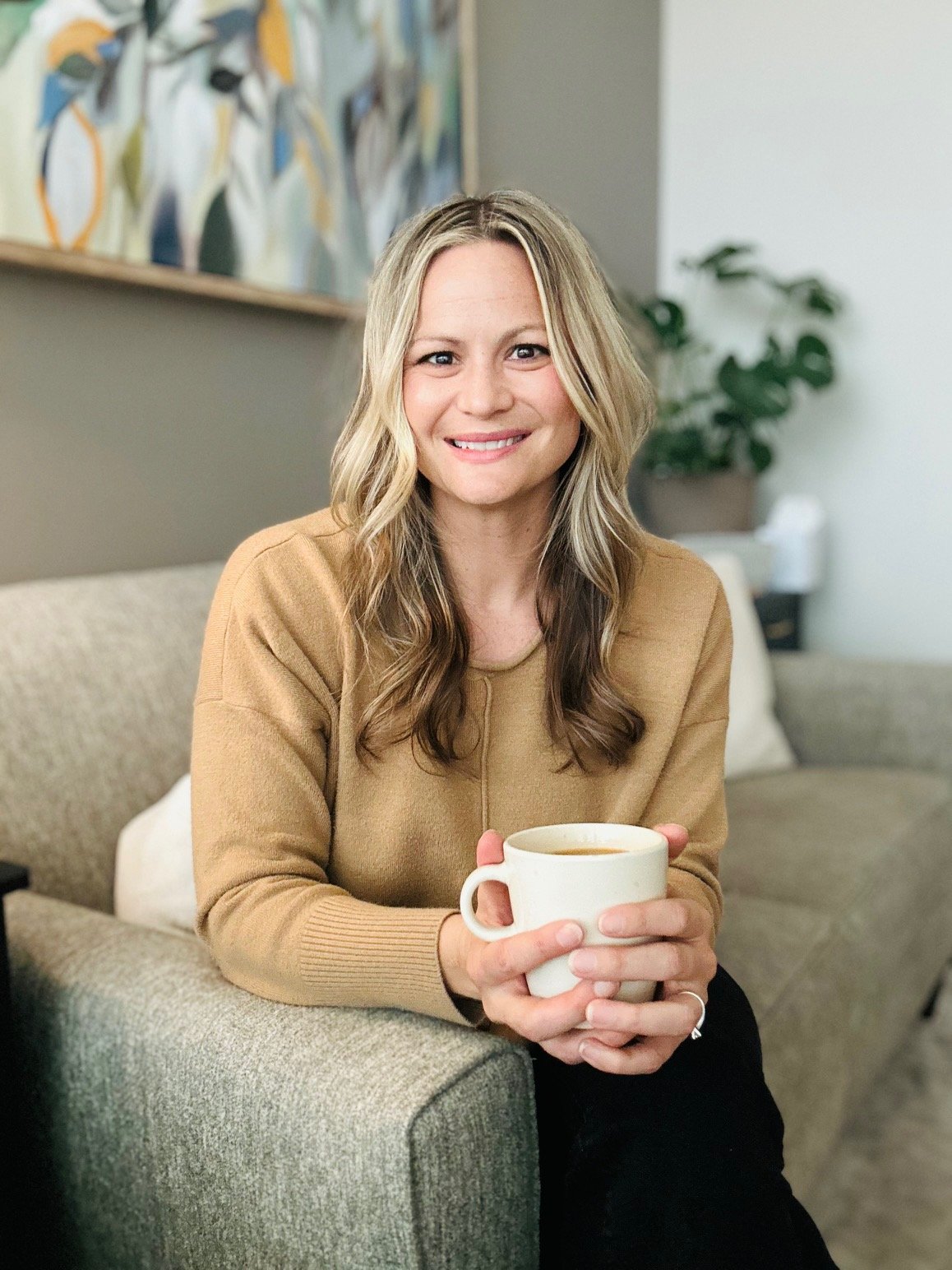 A woman with long blonde hair smiling while sitting on a beige sofa, holding a white mug of coffee, in a living room with a colorful painting and a green potted plant in the background.
