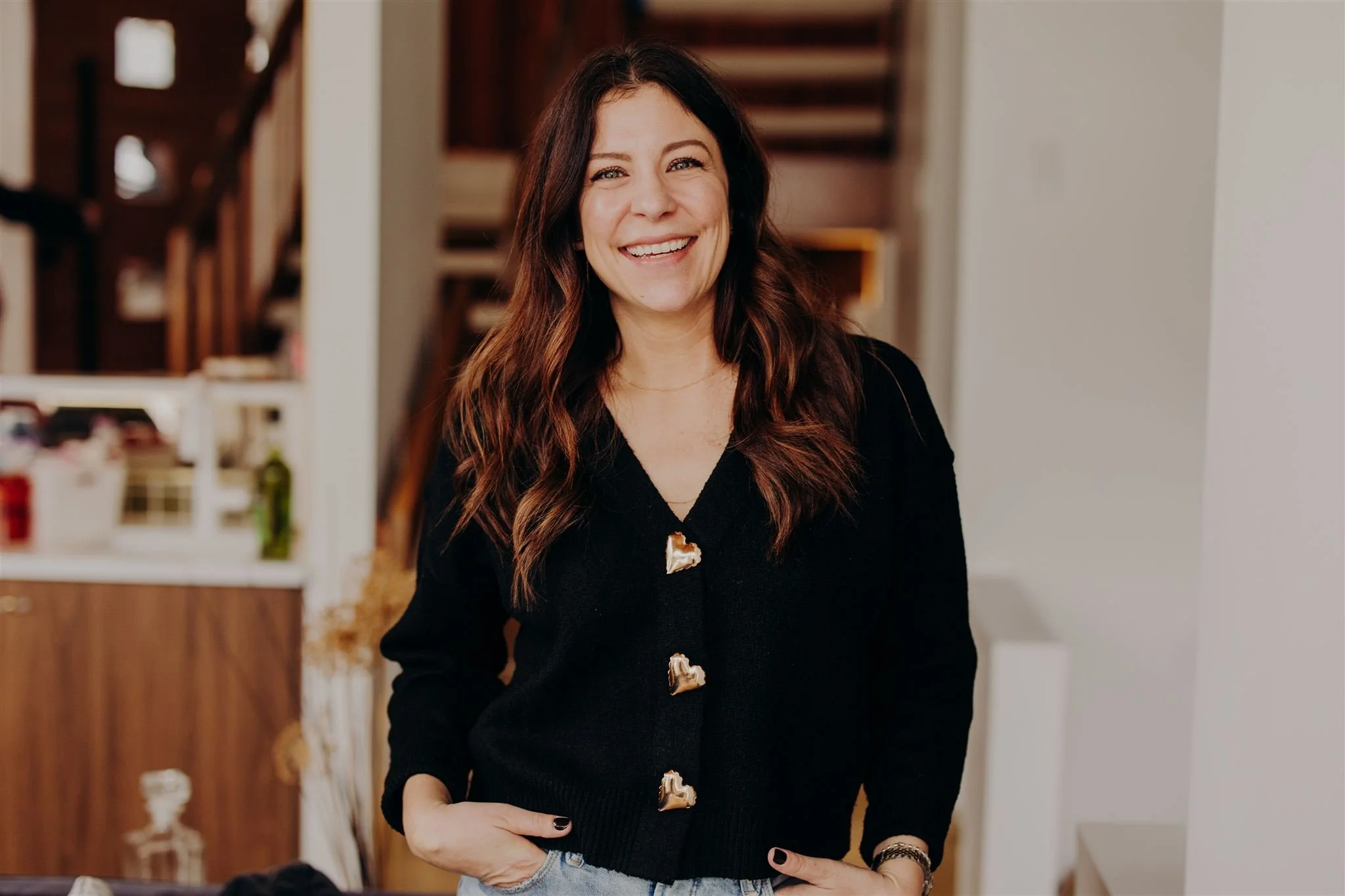 A woman with long, wavy brown hair and a big smile, standing indoors in a room with a staircase in the background, wearing a black sweater with shell-shaped buttons and light blue jeans.