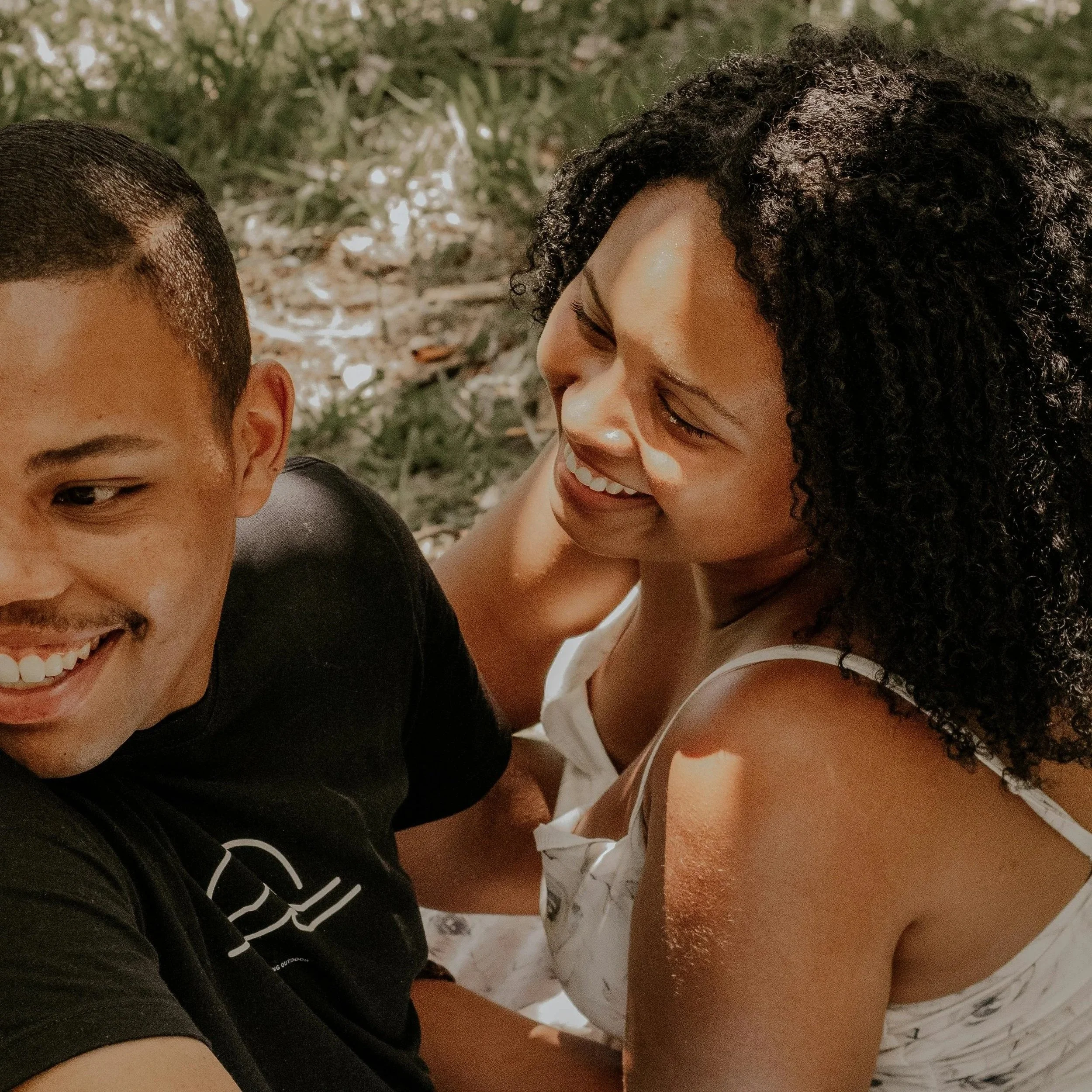 Close-up of a smiling woman with curly hair leaning in towards a young man with short hair, against a blurred background of Christmas tree branches with lights.