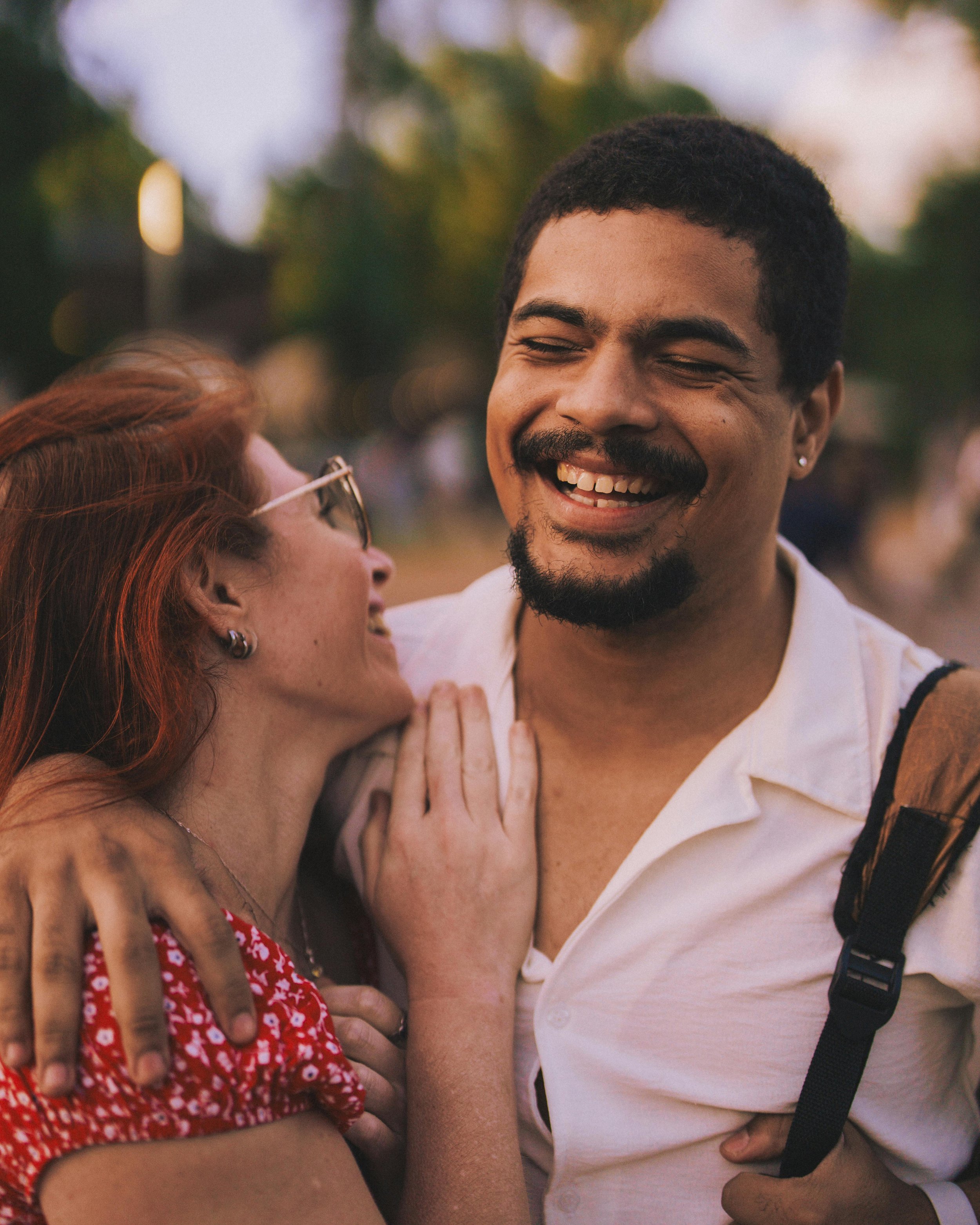 A close-up of a man with a mustache and beard smiling and looking at a woman with red hair and sunglasses, who is smiling back. They are in an outdoor setting with blurred trees and lights in the background.