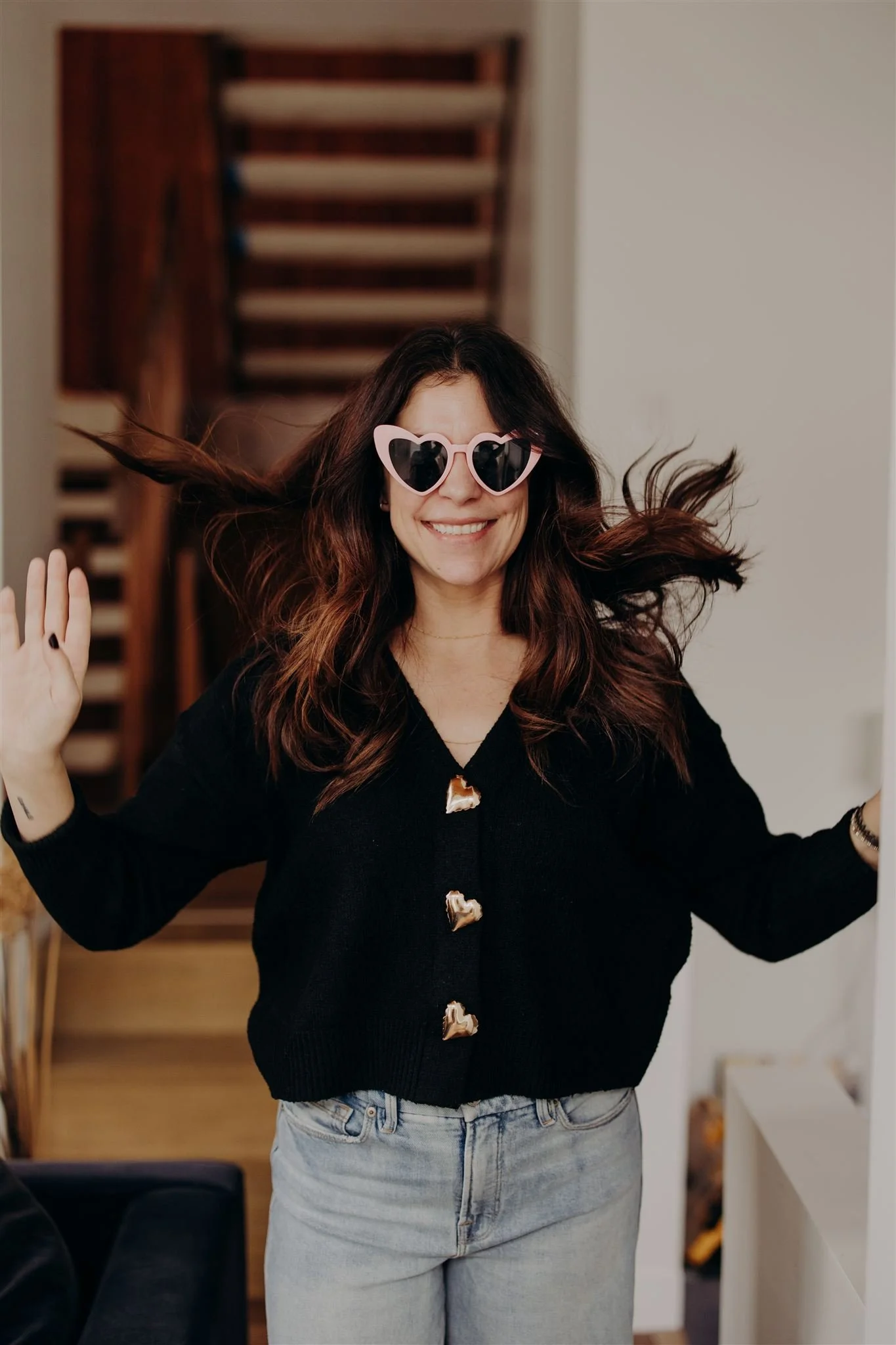 Woman with long wavy brown hair wearing heart-shaped sunglasses, smiling, wearing a black cardigan with heart-shaped buttons, and light-washed jeans, indoors with staircase in background.