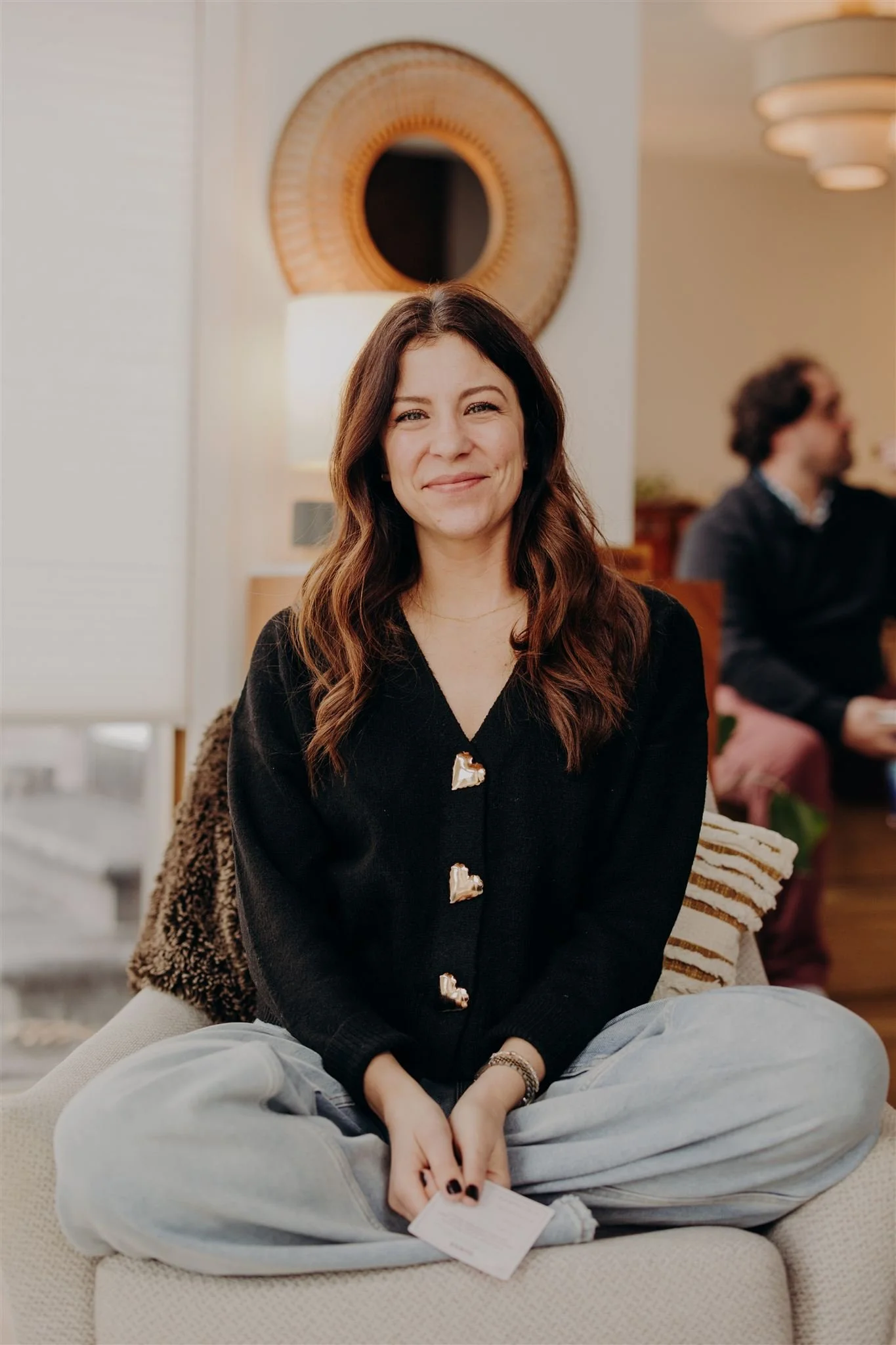 A woman with long brown hair sitting cross-legged on a beige cushioned seat, smiling at the camera, holding a small white card. She is wearing a black sweater with decorative buttons, light gray pants, and has a gold watch on her wrist. In the background, there is a blurred man with curly hair sitting on a chair, a wall with a circular mirror, and a lamp.