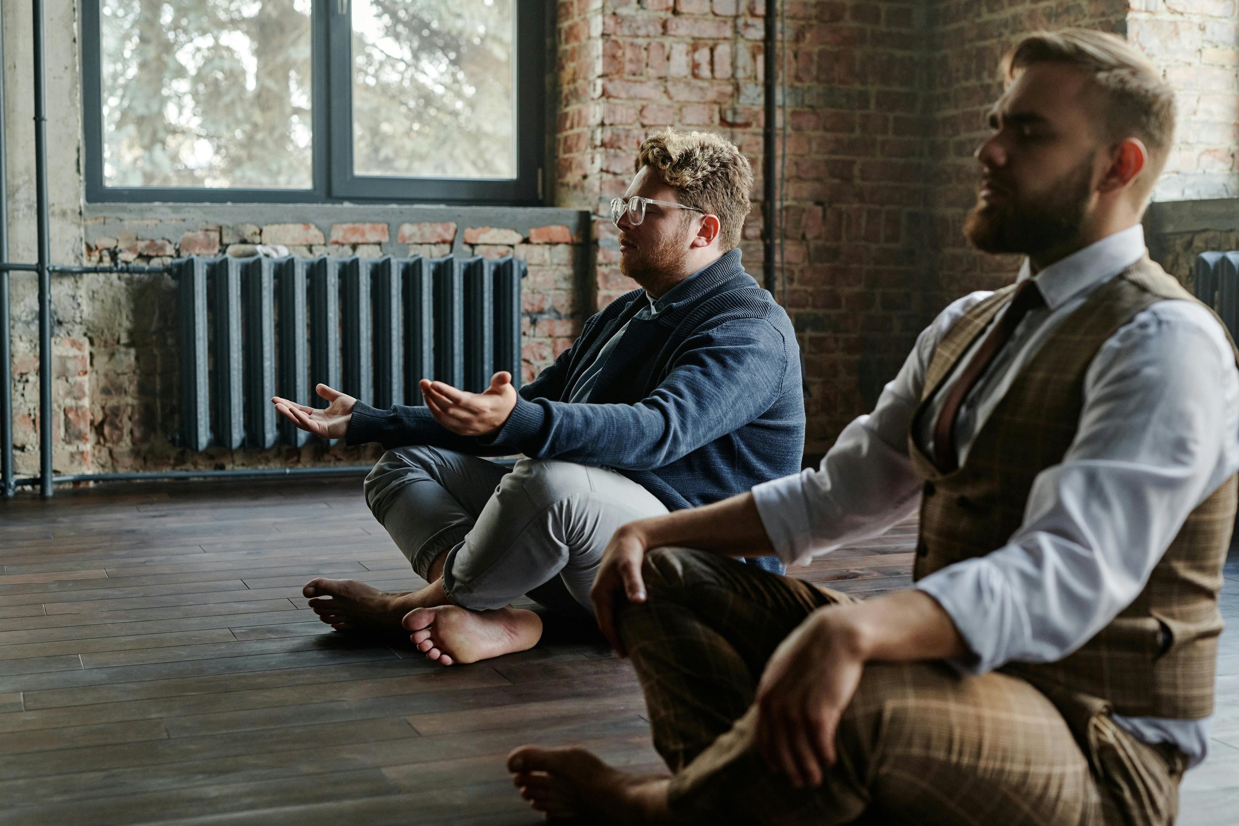 Two men sitting cross-legged on wooden floor, meditating in a room with brick walls and large windows, one wearing glasses and a dark blazer, the other with a beard and checkered vest.