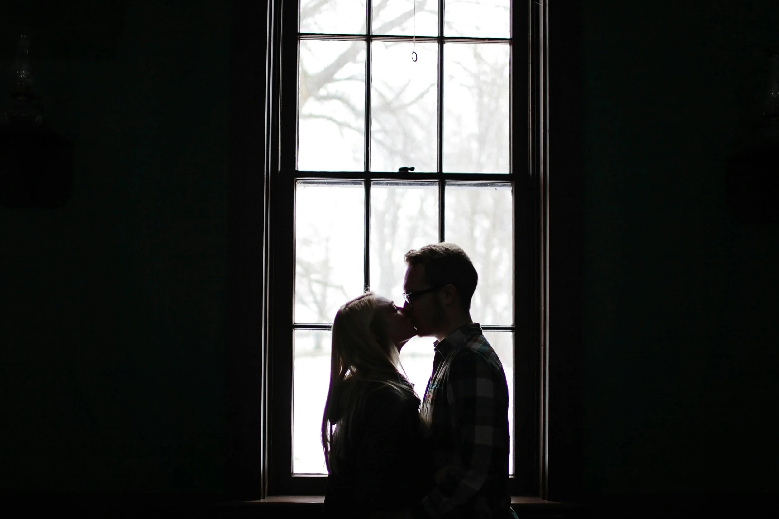 Silhouette of a couple kissing in front of a window with bare tree branches outside.