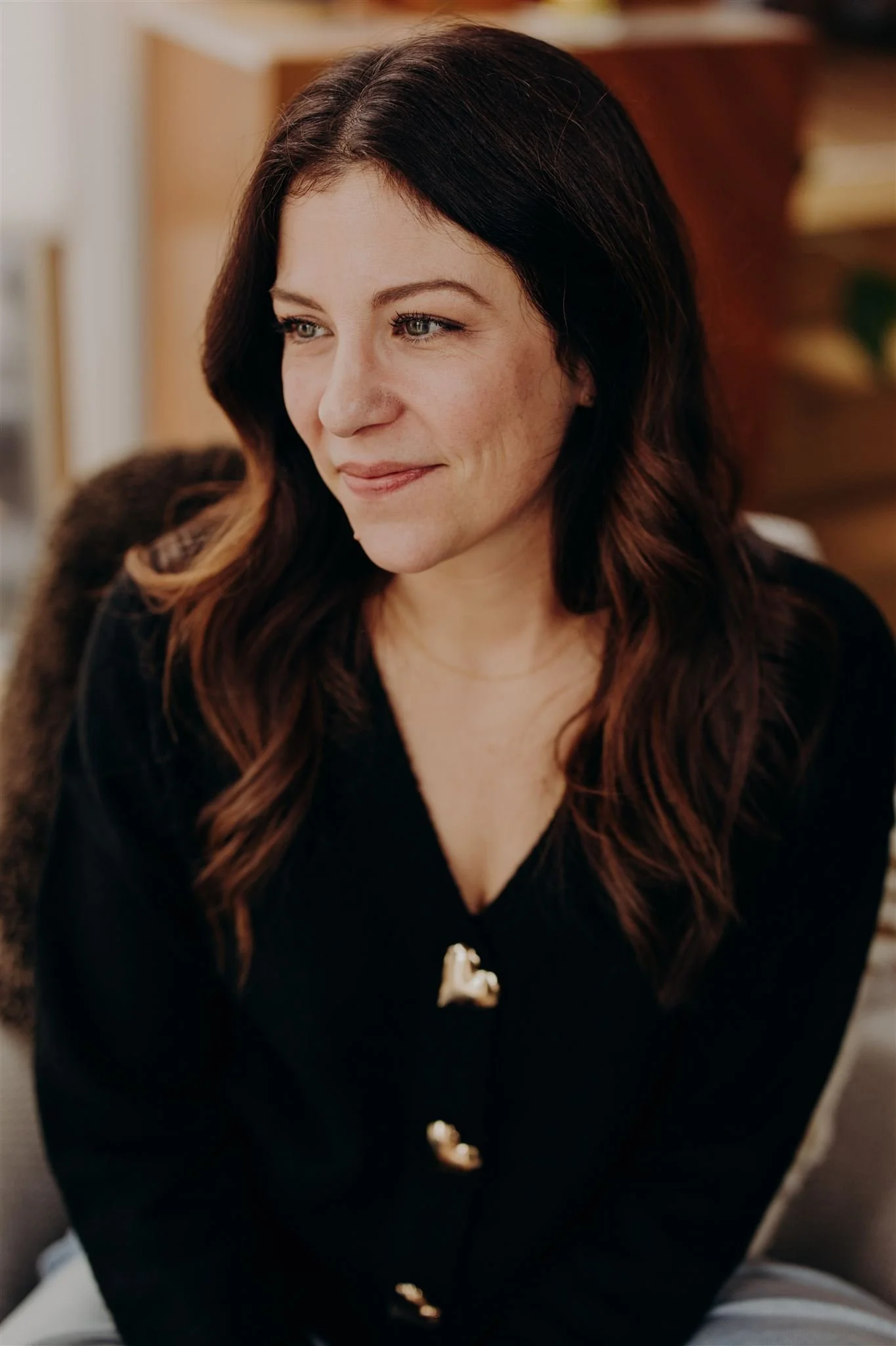 A woman with long, wavy, dark brown hair and light skin, smiling softly while sitting indoors, wearing a black top with a gold pin.