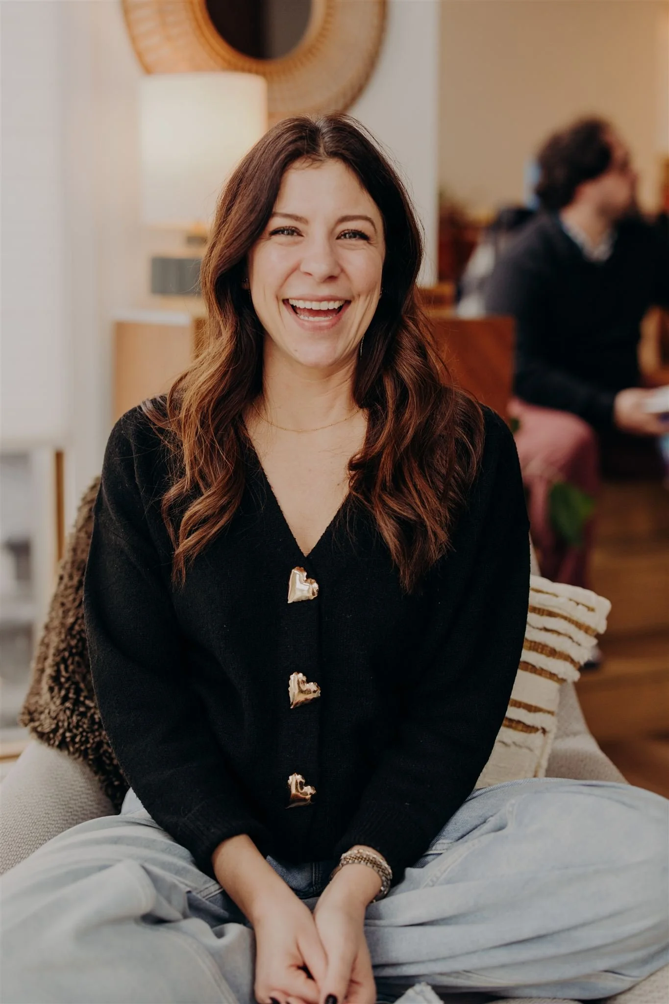 A woman with long brown hair smiling and sitting cross-legged on a couch in a cozy living room.