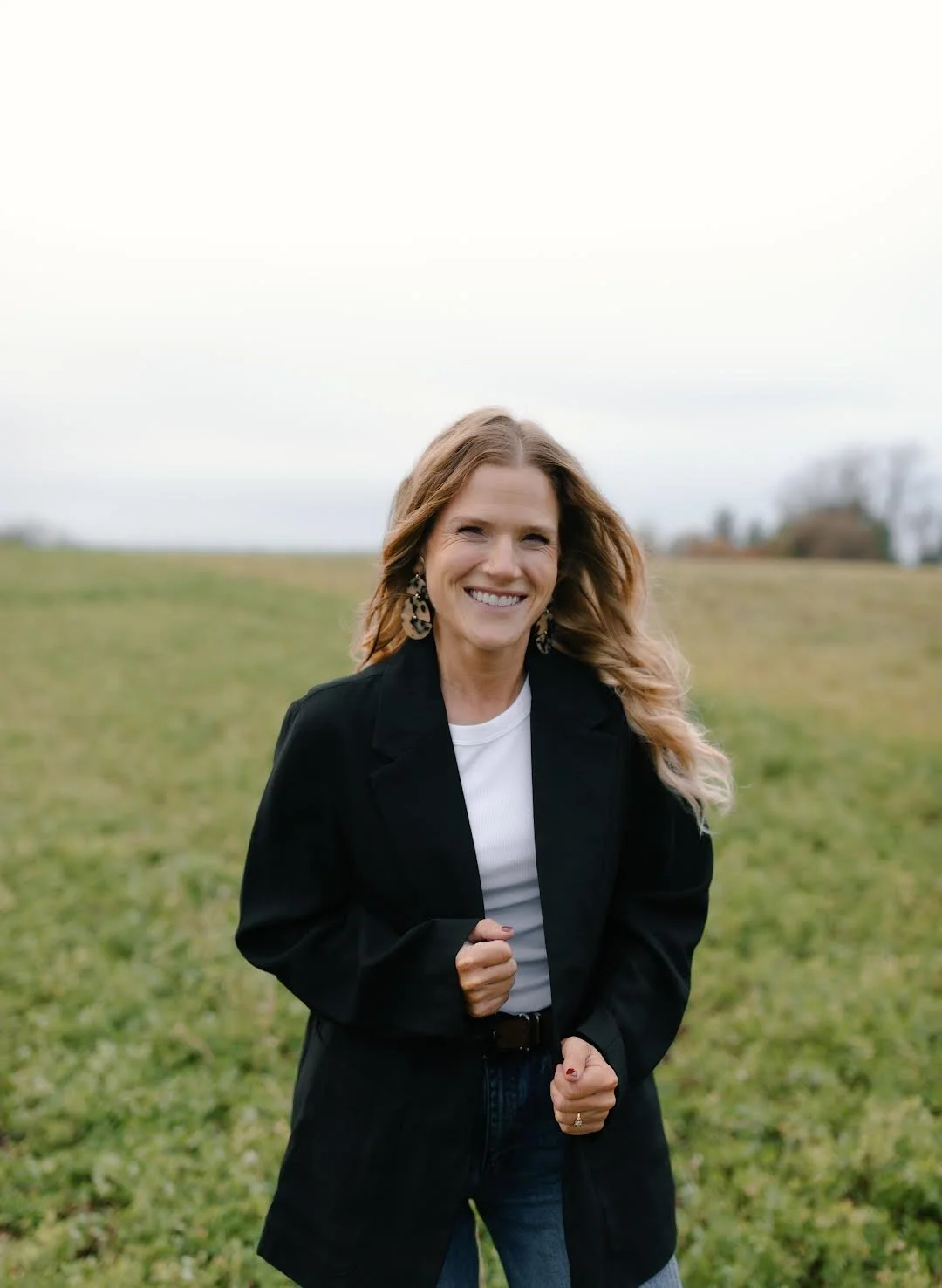A woman with light skin and wavy, shoulder-length hair smiling outdoors in a grassy field on a cloudy day.