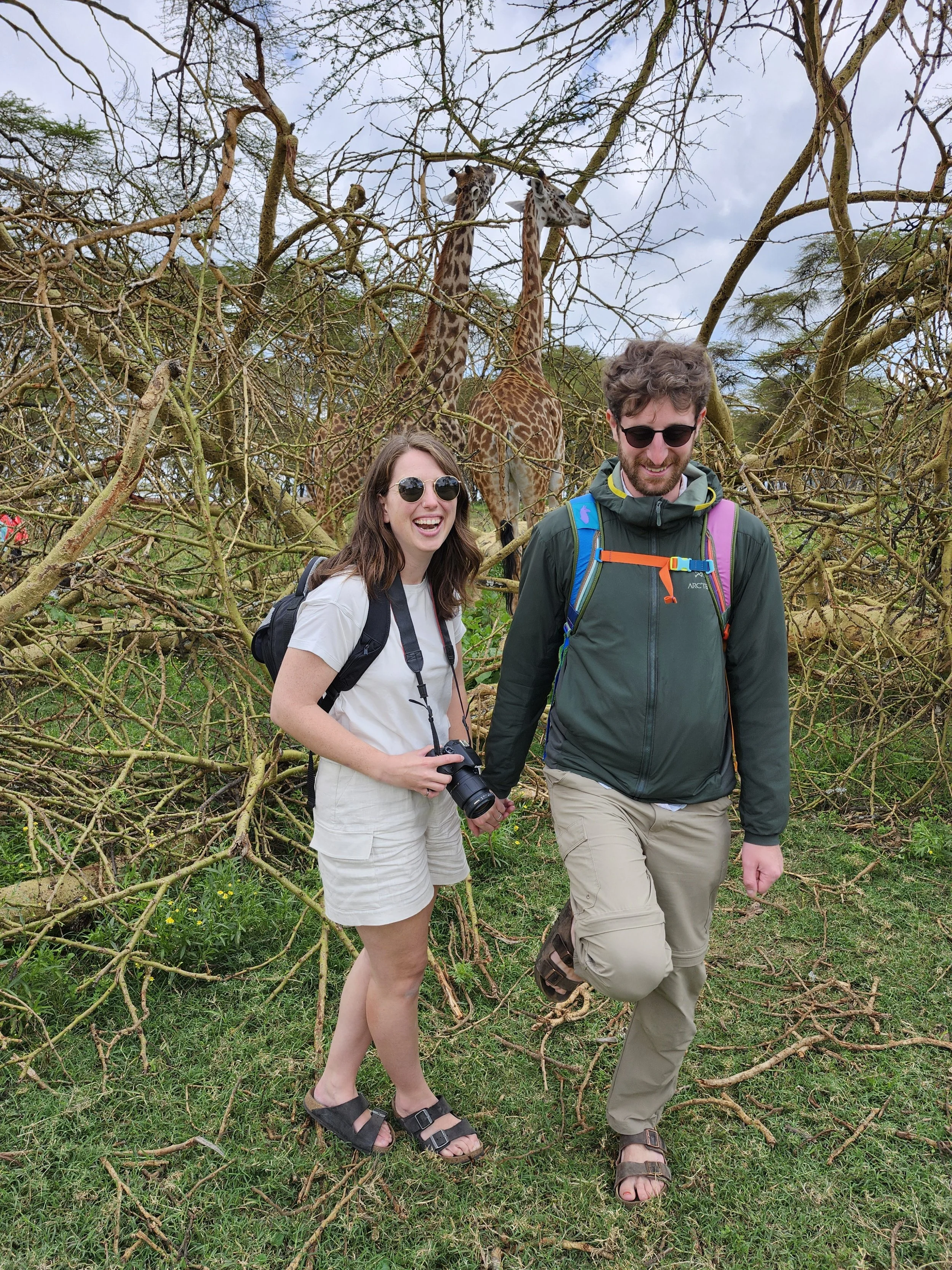 Two people, a woman and a man, standing in a grassy area with branches and leafless trees behind them. They are smiling and holding hands. The woman has long brown hair, sunglasses, a white t-shirt, beige shorts, and sandals. The man has curly hair, sunglasses, a dark jacket, beige pants, and sandals. They are in front of two giraffes standing in a tree.