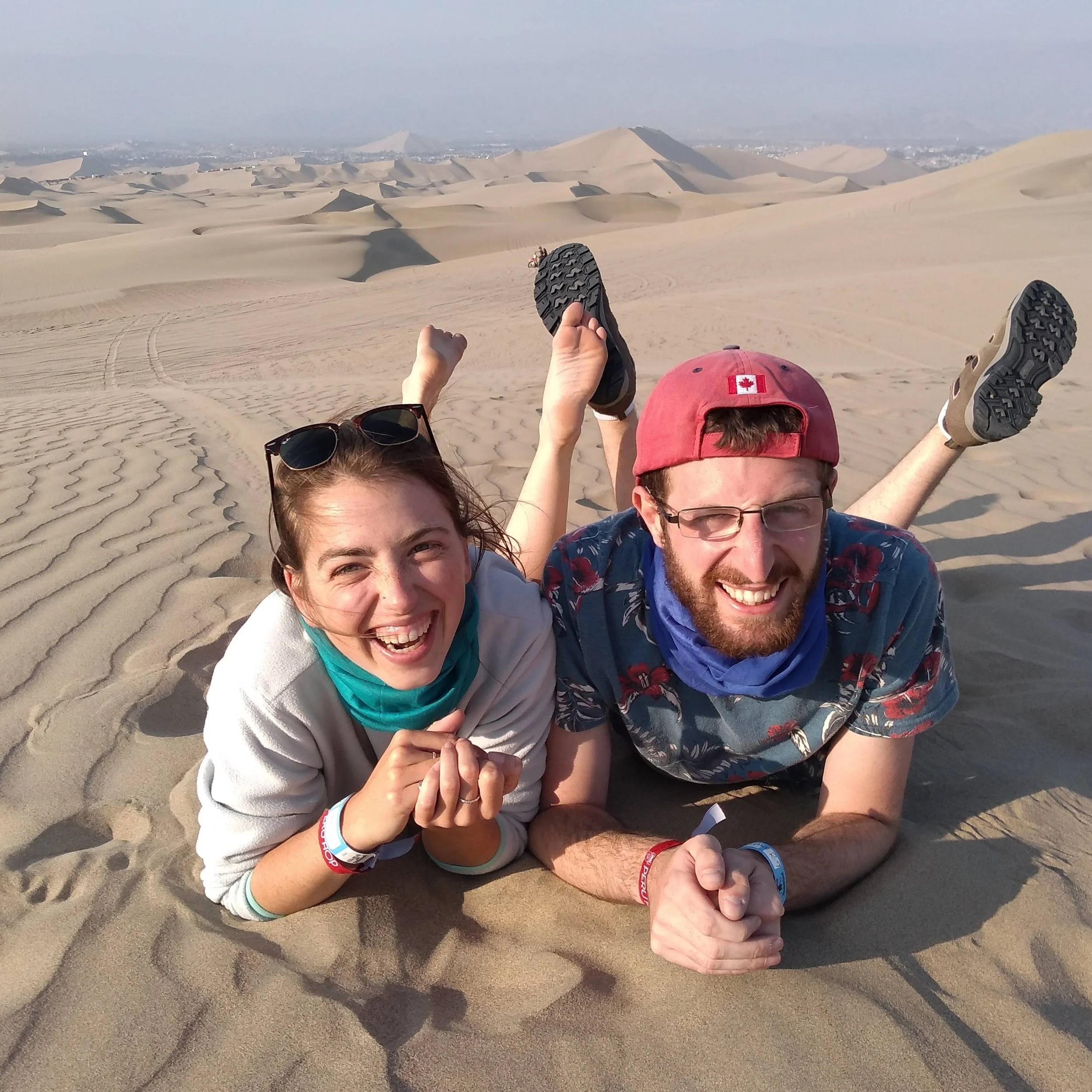 People lying in a desert with sand dunes in the background, some with their legs and arms raised, wearing casual clothing and wristbands, enjoying a sunny day.
