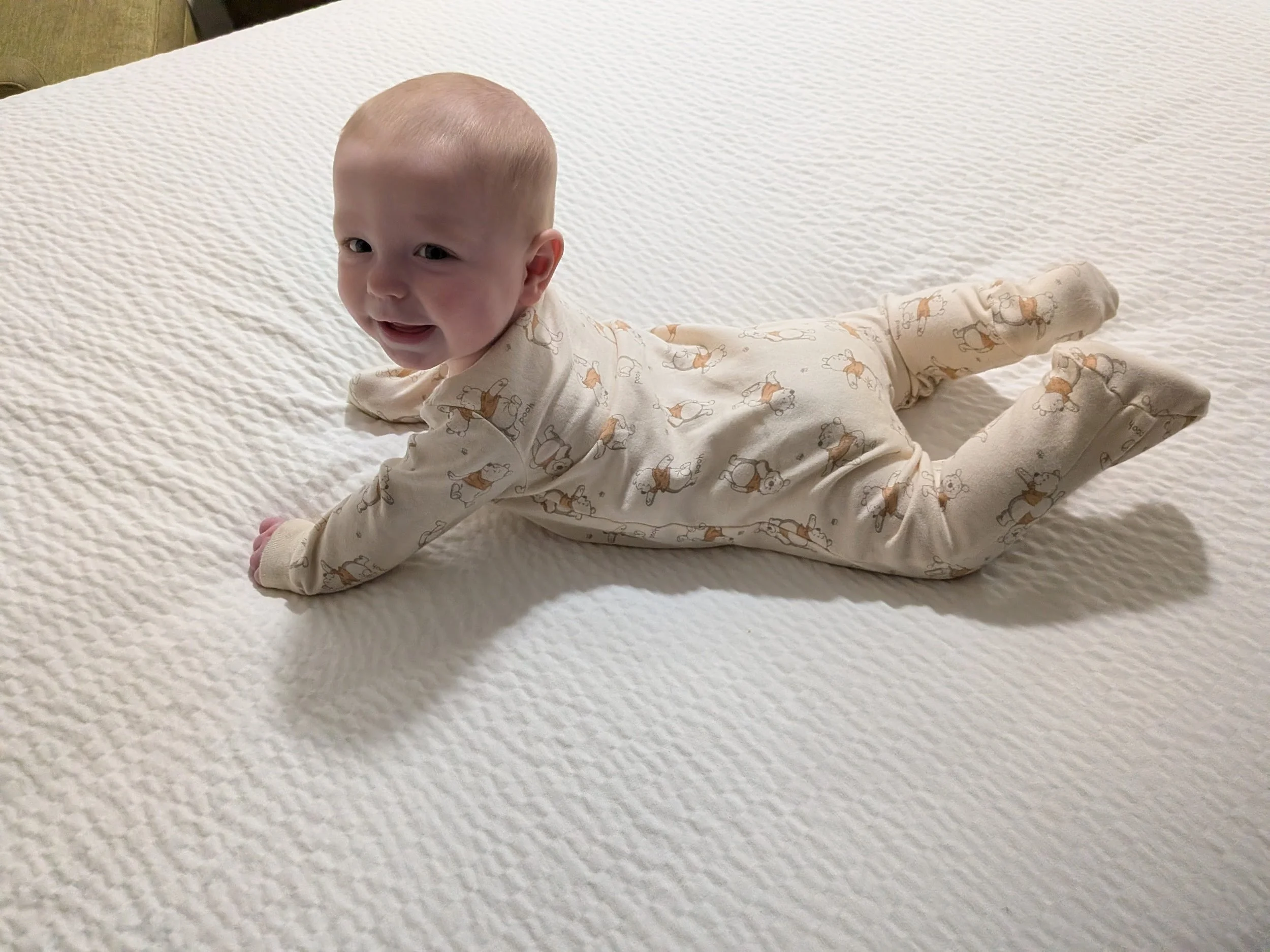A baby lying on a white textured bedspread, looking upward and smiling with an open mouth, wearing cream-colored pajamas with a bear pattern.