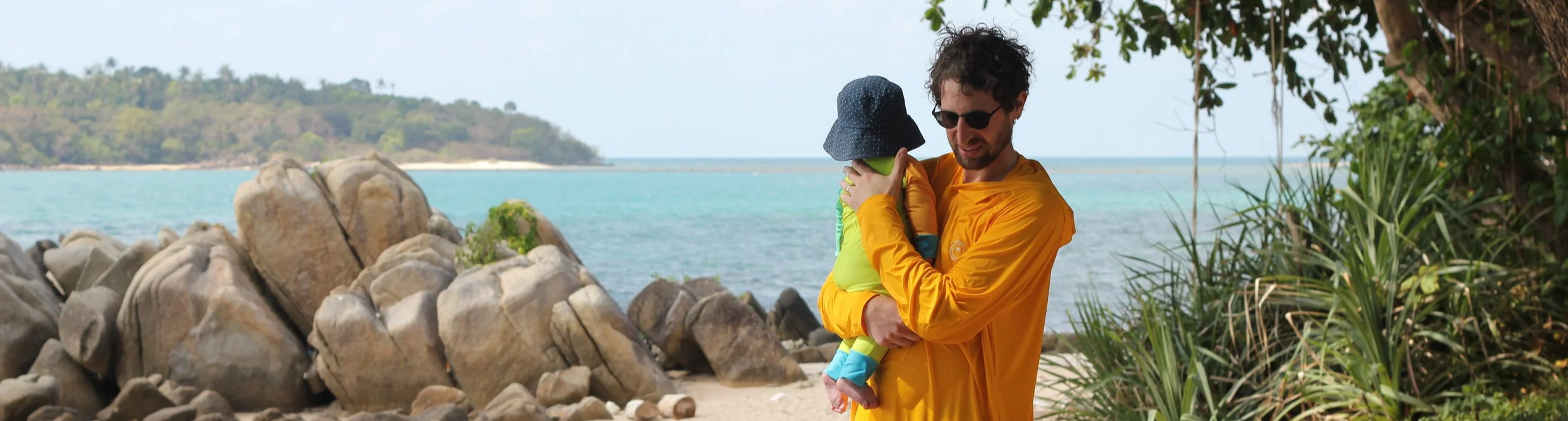 A man in an orange sun shirt holding a small child in a yellow sunsuit and hat on a beach with rocks, trees, and water in the background.