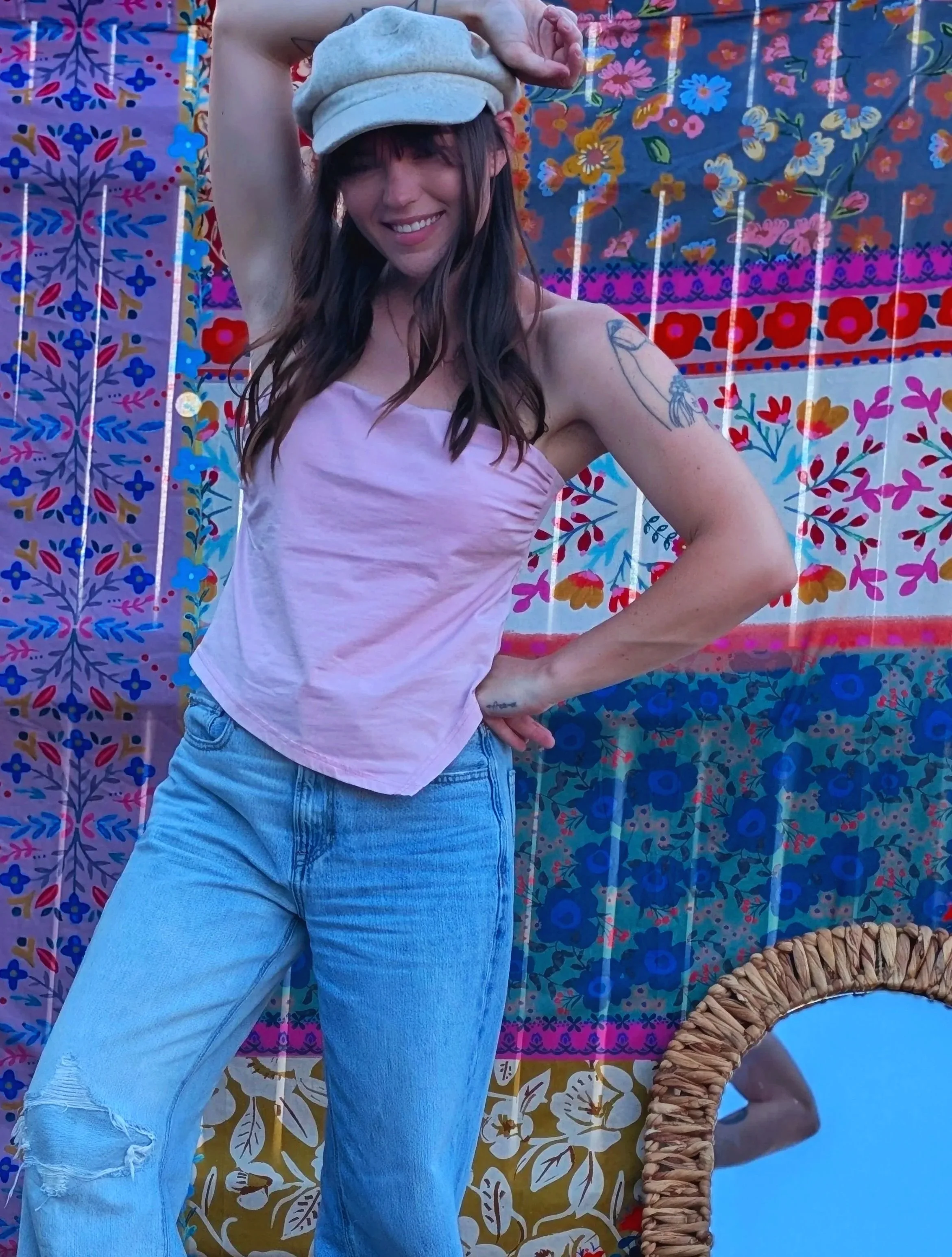 A woman with long dark hair wearing a tan cap, a purple tank top, and light blue ripped jeans, posing with her hand on her hip in front of a colorful floral patterned backdrop.