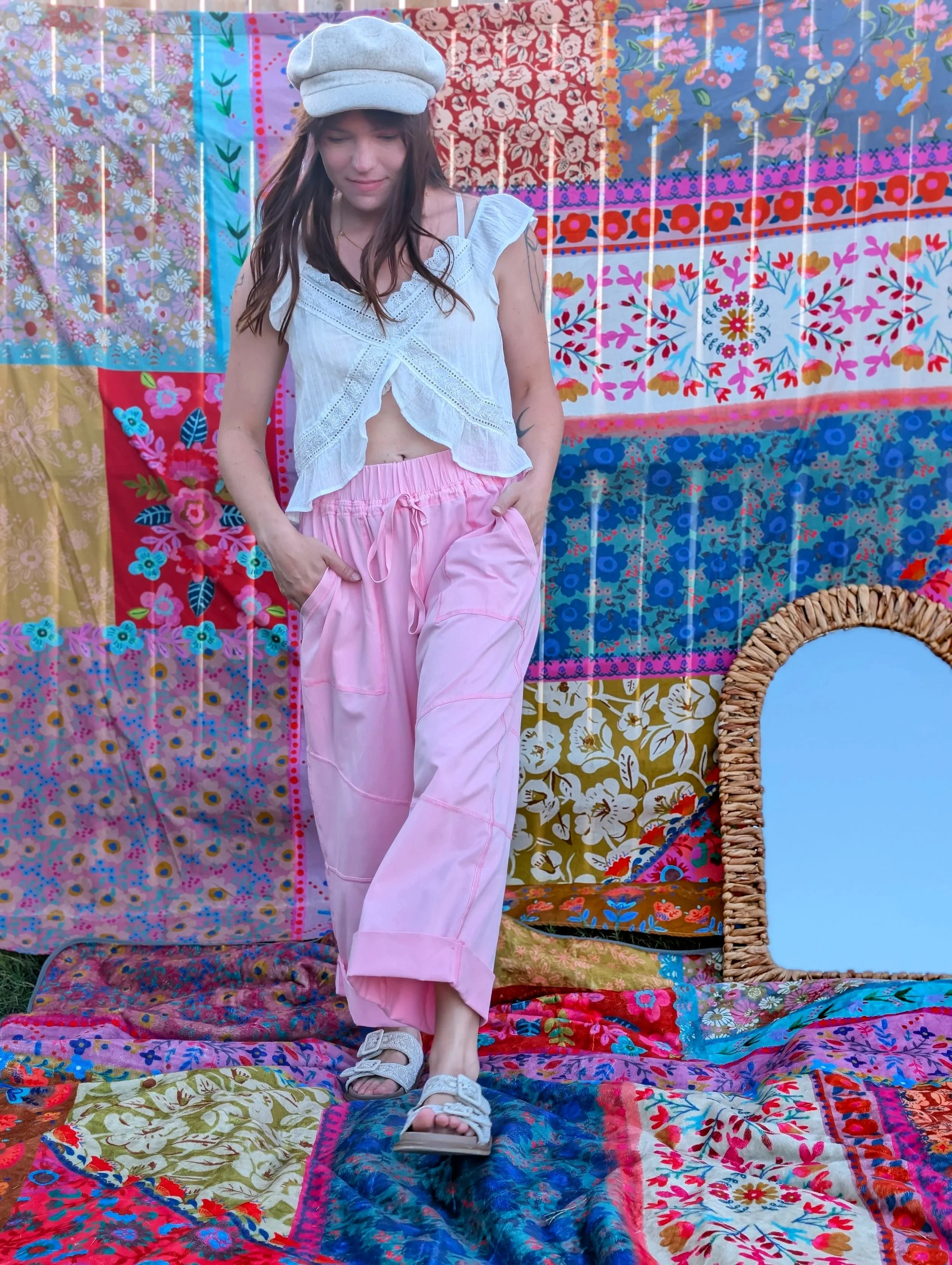 A young woman with long brown hair is standing on a colorful patchwork quilt, wearing a white sleeveless top, pink pants, white sandals, and a grey cap. She is looking down slightly and has one hand in her pocket. The background is a vibrant patchwor