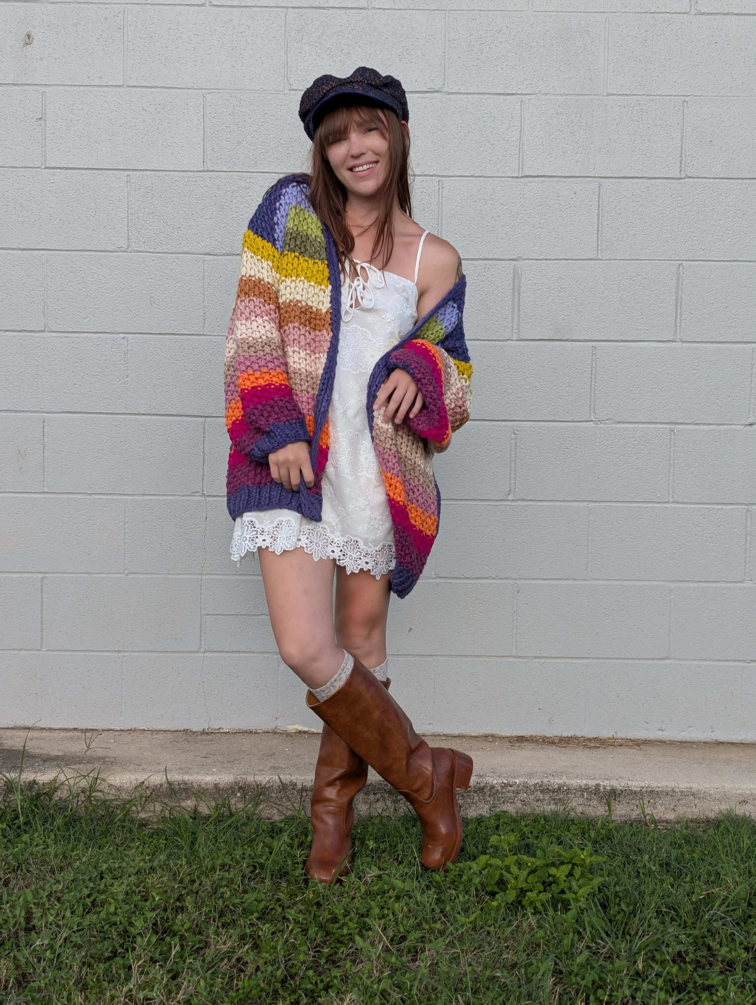 A woman with brown hair, wearing a white lace dress, colorful striped knit cardigan, brown knee-high boots, and a newsboy cap, standing outdoors against a gray brick wall, smiling, with grass underfoot.