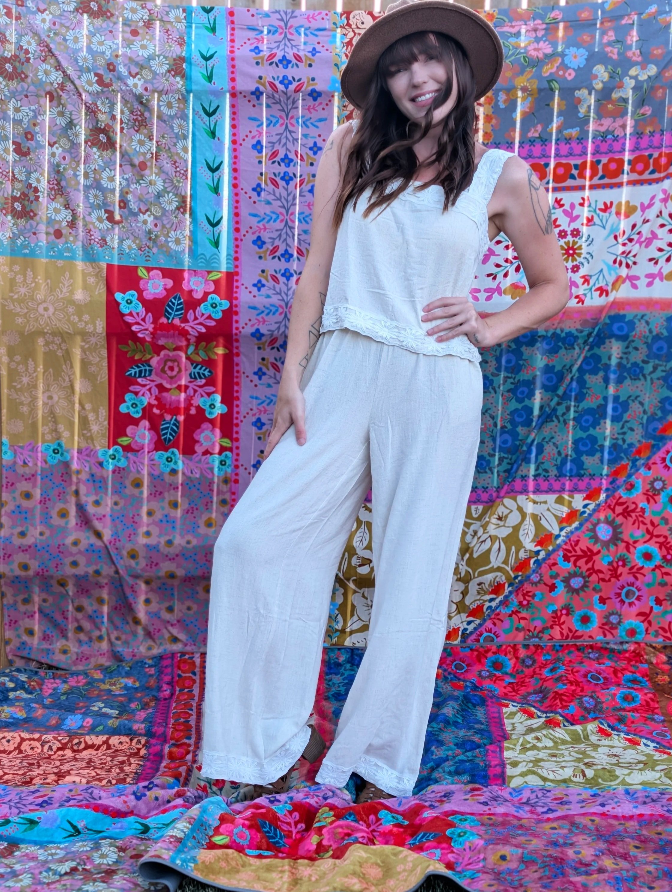 A woman wearing a white sleeveless top and wide-legged white pants, with a wide-brimmed brown hat, standing in front of a colorful patchwork quilt display. She has a smile, dark brown hair with bangs, and tattoos on her arm.