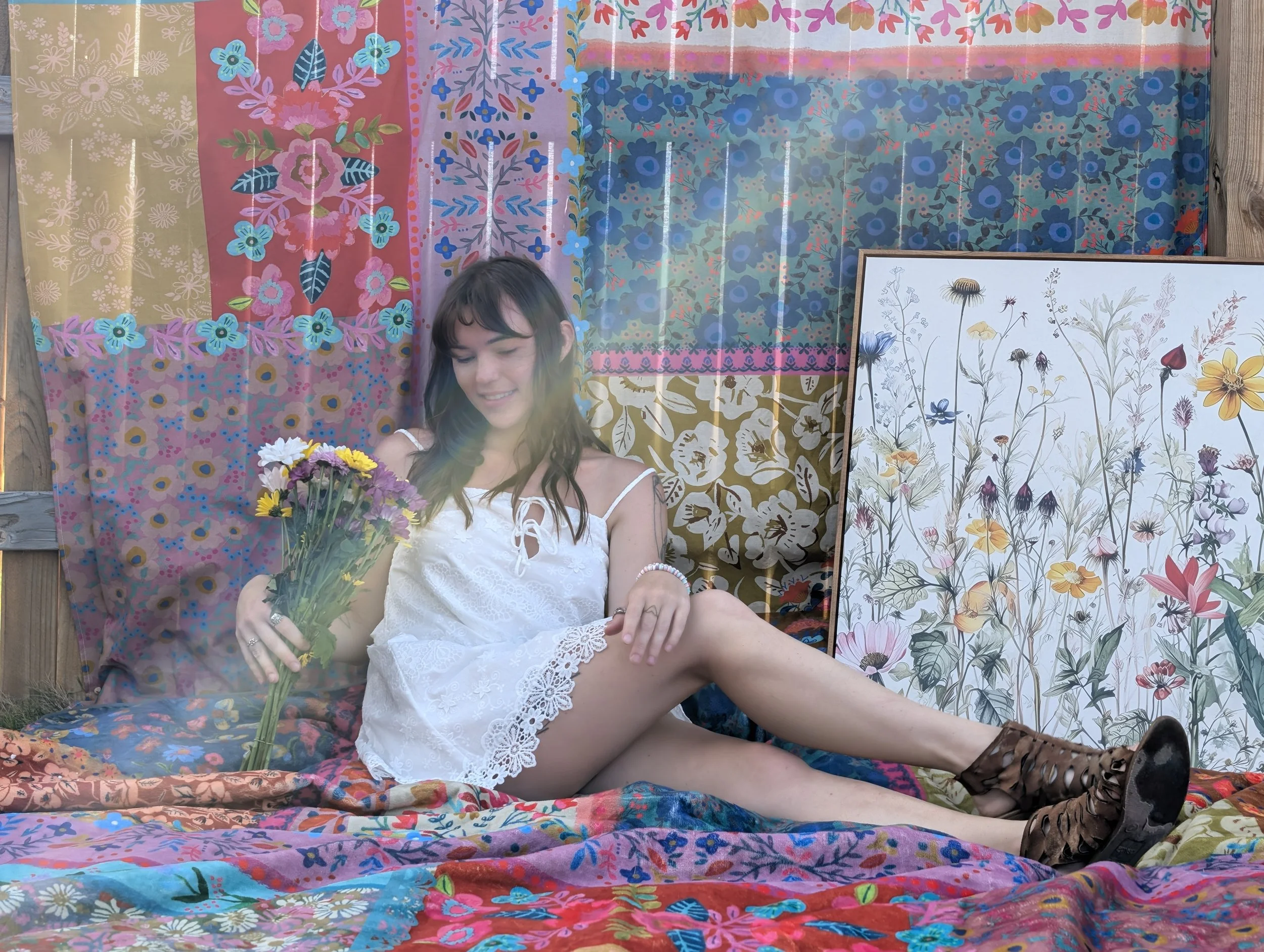 A young woman with dark, wavy hair sitting on a colorful bed with floral sheets and blankets, holding a bouquet of flowers, outdoors in front of a vibrant, patterned backdrop with floral designs. She is wearing a white lace dress, smiling with her ey