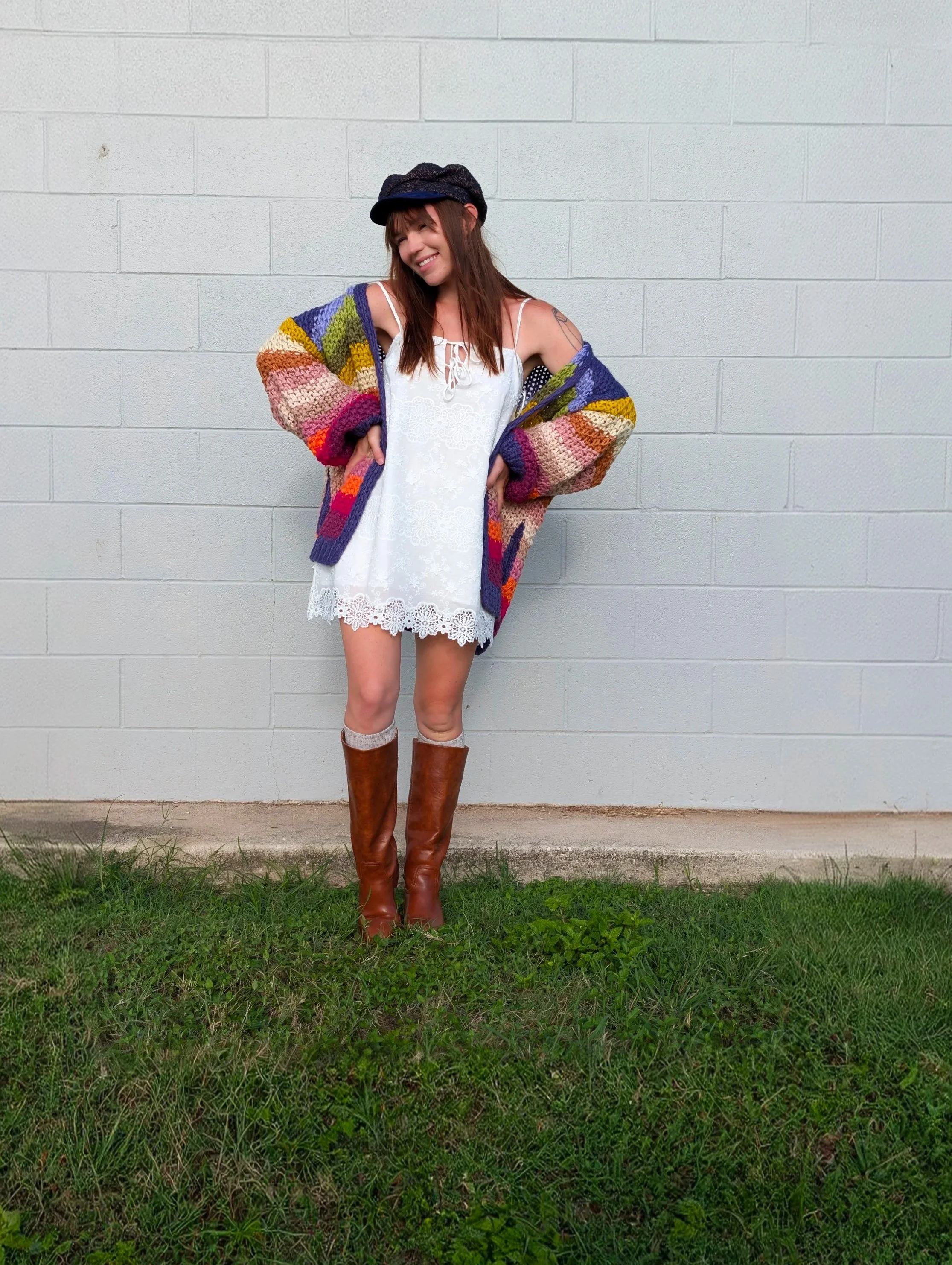A woman with light skin and brown hair standing outdoors against a gray brick wall, smiling and looking down. She is wearing a black hat, a colorful striped cardigan, a white lace dress, and brown tall boots.