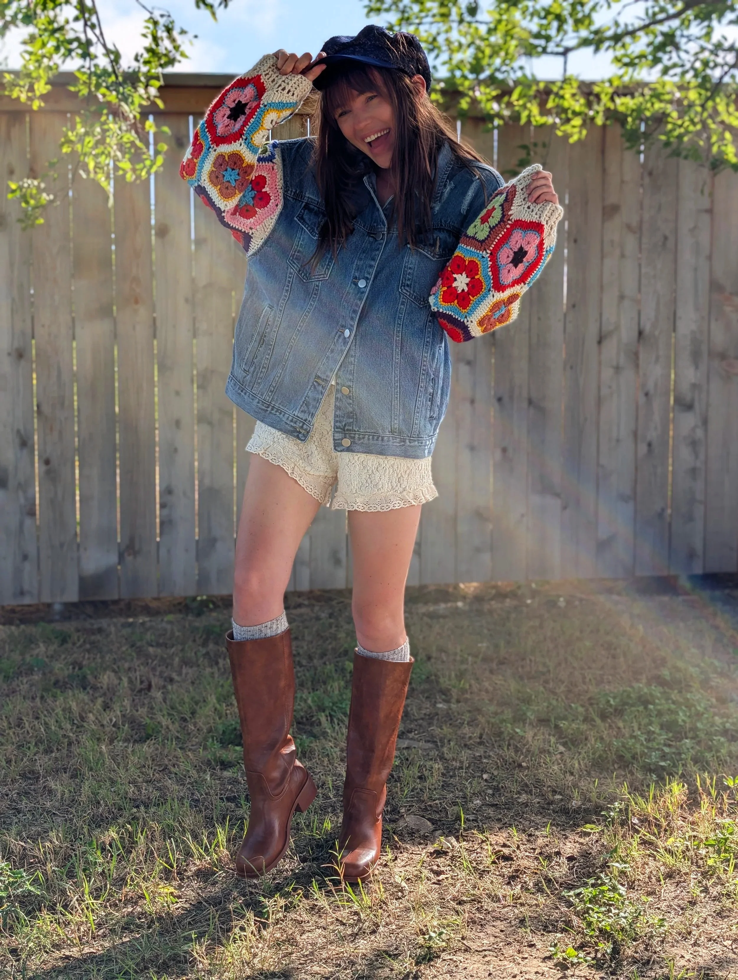 A woman standing outdoors on grass, smiling, wearing a denim jacket with colorful crocheted sleeves, cream lace shorts, brown knee-high boots, and a black cap, with a wooden fence and trees in the background.