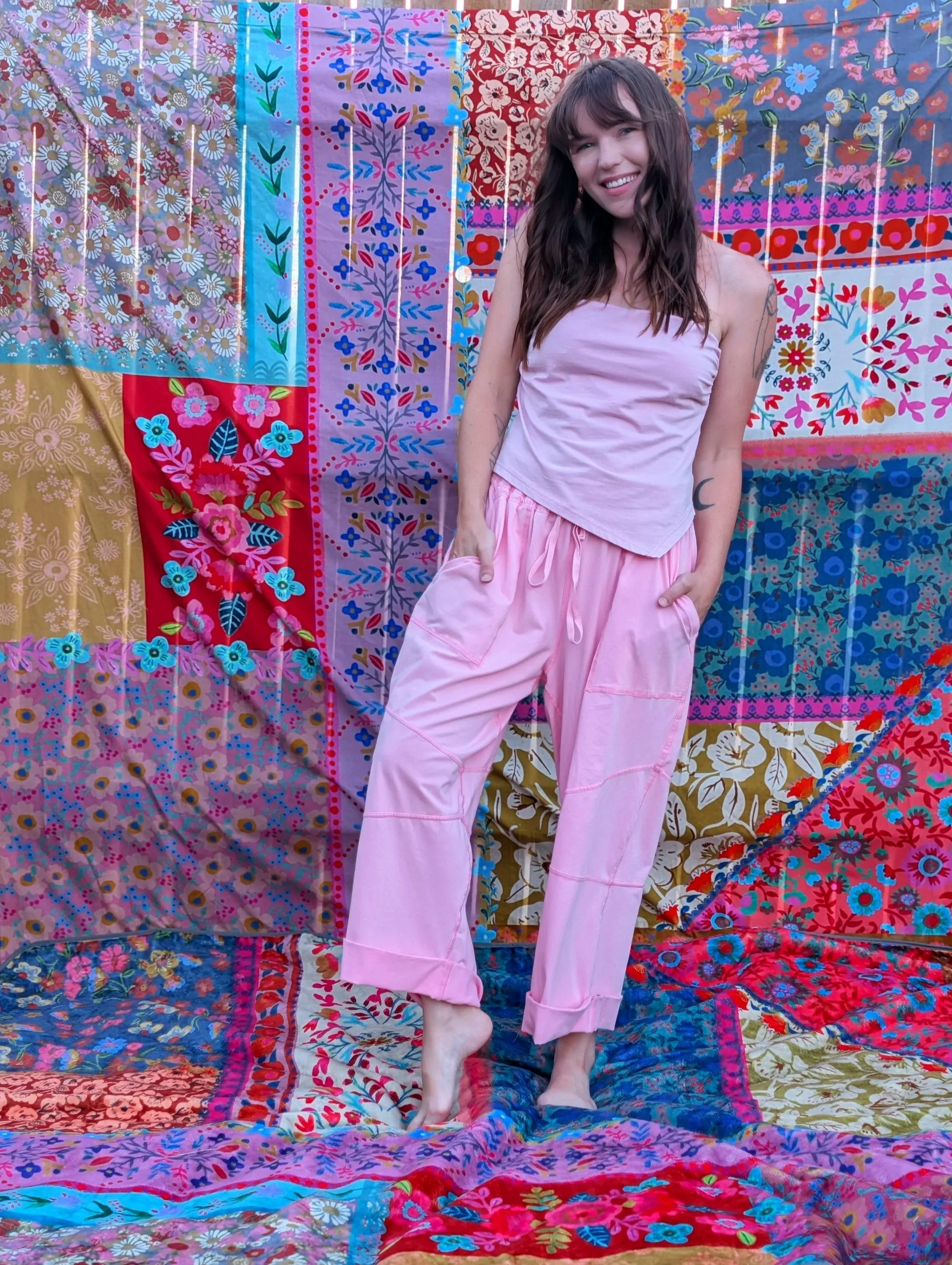 A woman with long wavy brown hair and tattoos on her arm stands barefoot in front of a colorful patchwork quilt with floral and abstract patterns. She is wearing pink pajama pants and a light pink tank top, smiling at the camera.