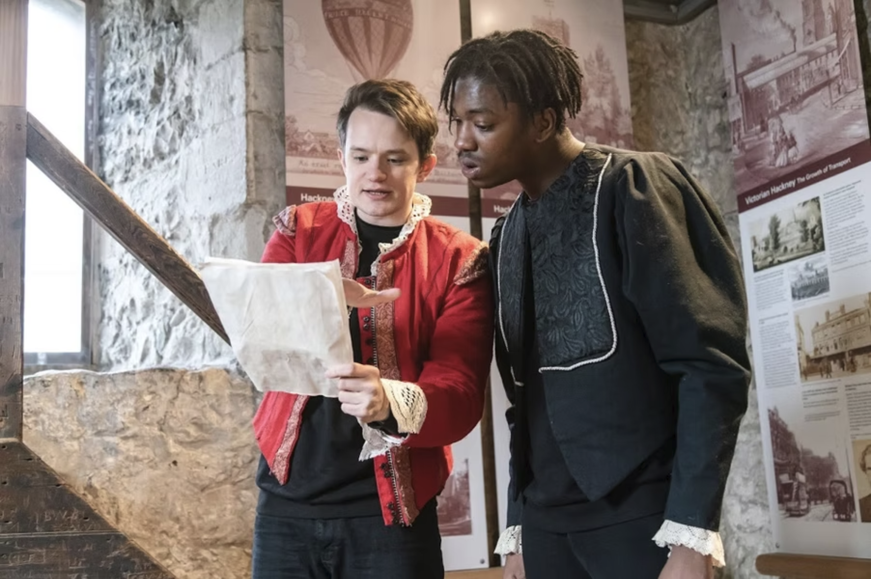 Two young men looking at a map inside a stone-walled museum or historical site. One wears a red jacket with lace details, the other a dark jacket with lace sleeves. Posters with historical images are in the background.