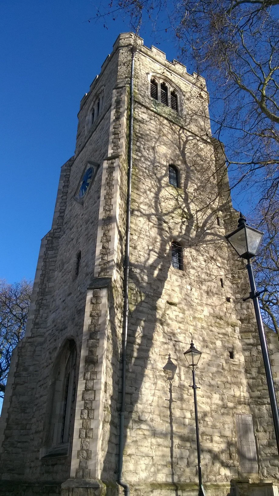 A tall stone church tower with small windows, a clock, and a trees casting shadows on its surface, under a clear blue sky.