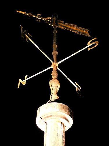 A weather vane shaped like a gold-colored arrow with a decorative tail, mounted on top of a stone column, pointing in multiple directions against a black background.