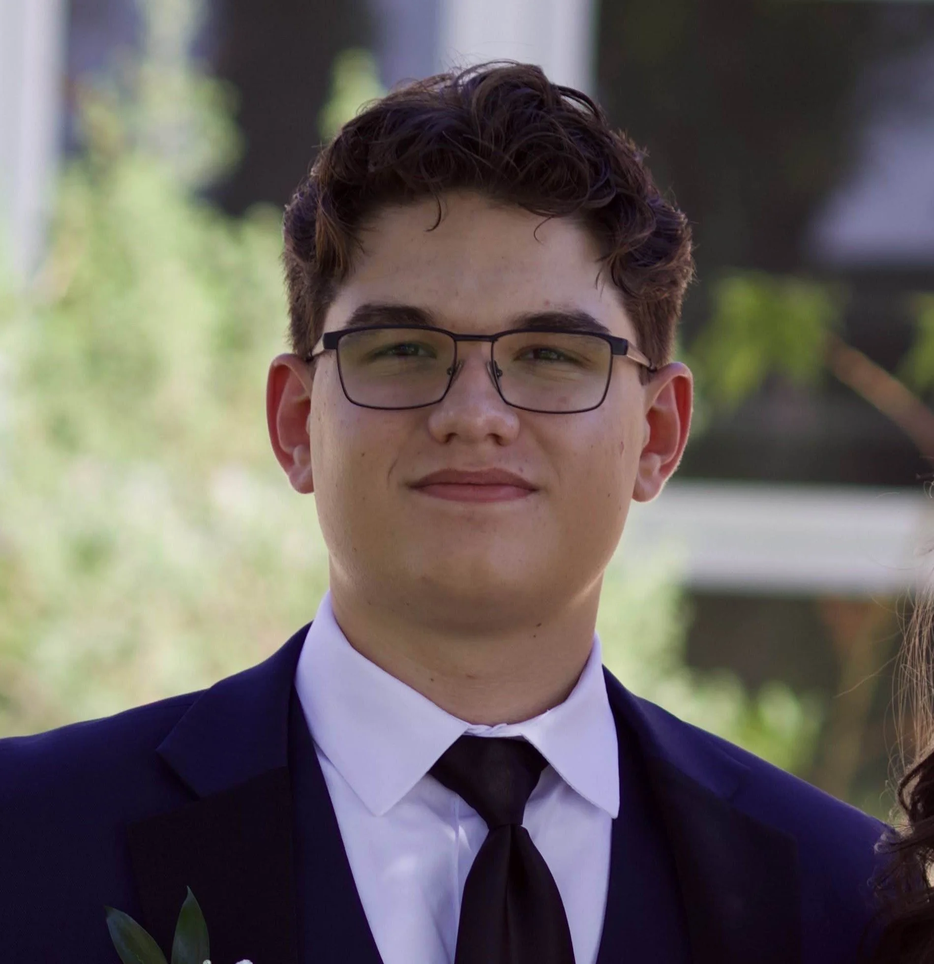 Close-up of Tristen Silva in formal attire, wearing glasses, with a blurred outdoor background.