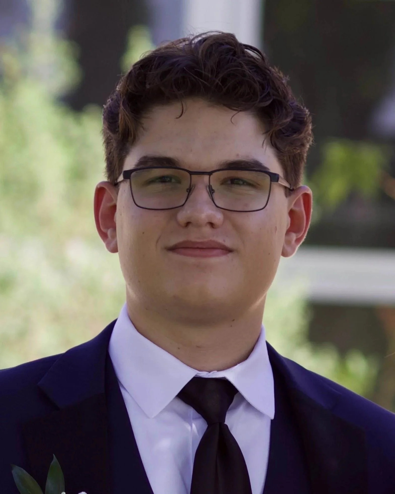 Close-up of a young man wearing glasses, a white shirt, a black tie, and a dark suit jacket, outdoors with a blurred background of trees and greenery.