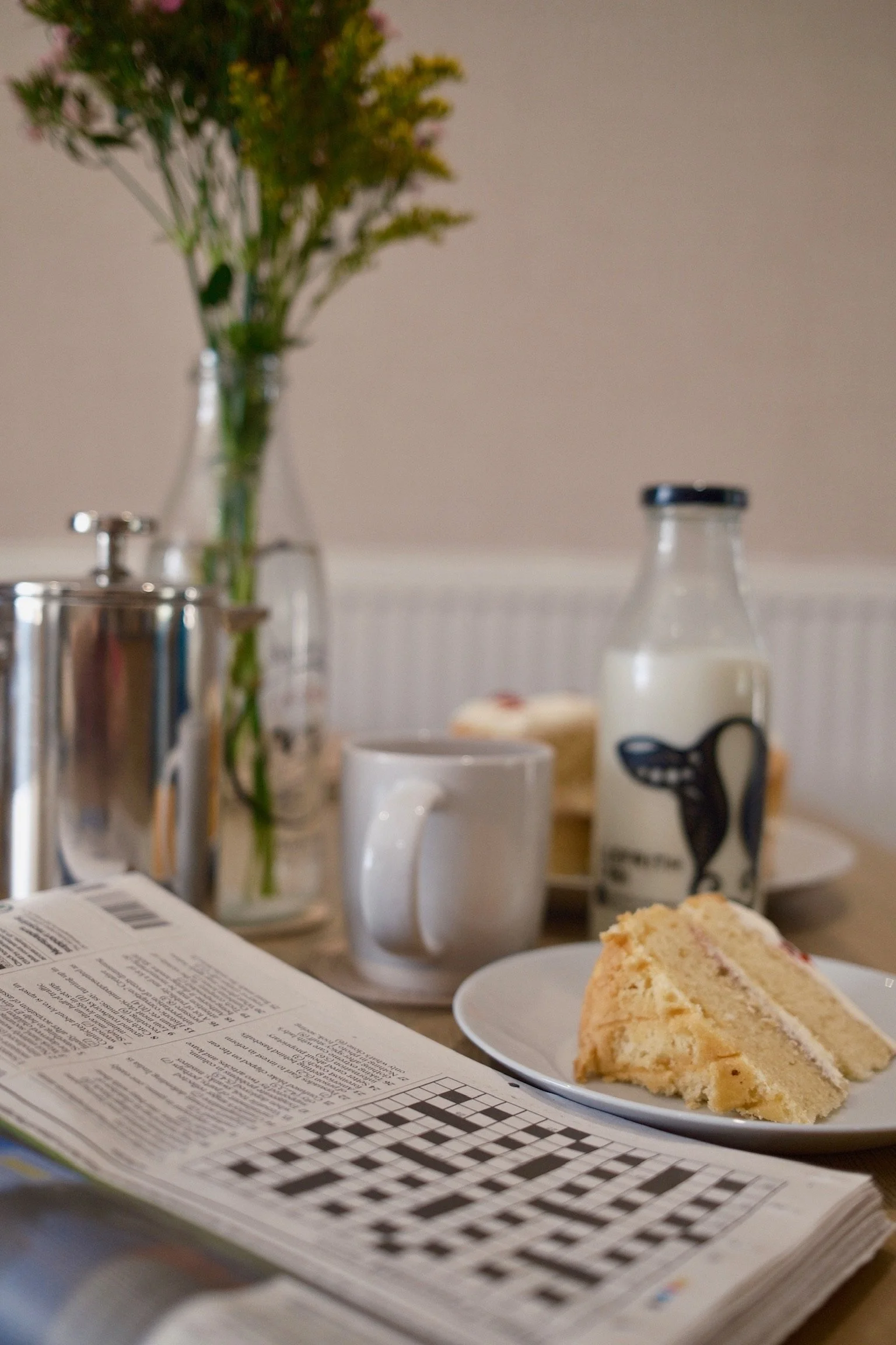 A partially eaten slice of cake on a white plate, a crossword puzzle book, a white mug, a bottle of milk, a potted plant, and a newspaper on a table.