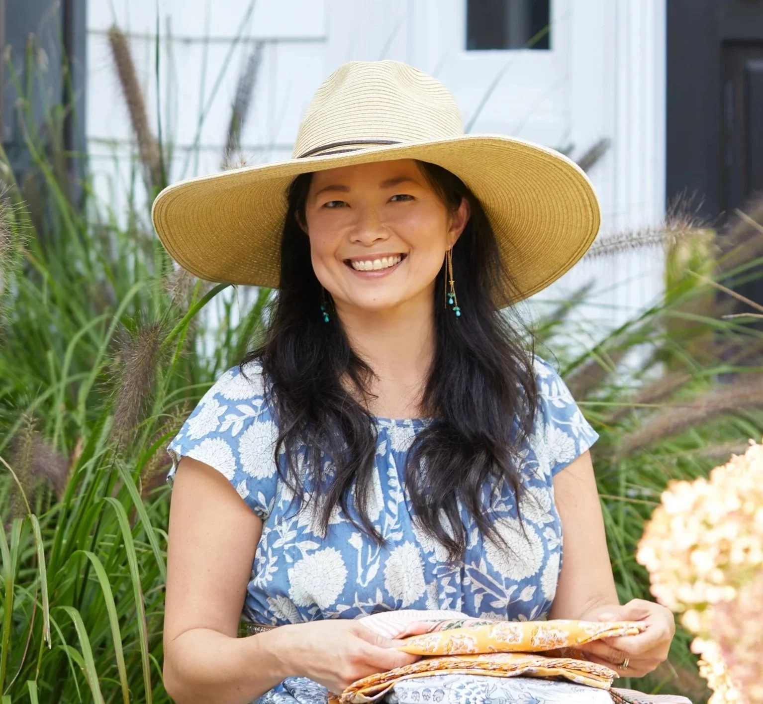 Woman in a blue and white floral dress with long dark hair, wearing a straw hat and turquoise earrings, sitting outdoors surrounded by greenery.